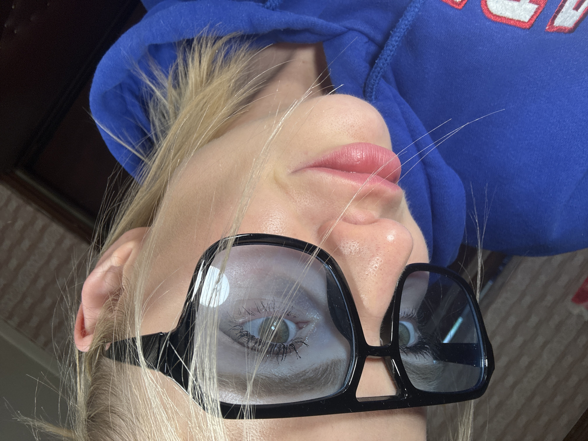 A close-up, slightly distorted, angled shot shows a fair-skinned person with blonde hair wearing large, black-framed glasses. The lenses of the glasses reflect light. The person is also wearing a blue hooded sweatshirt with red and white lettering partially visible. The background appears to be indistinct and out of focus. There is no discernible evidence indicating that this image was taken in Saint Petersburg, Russia.