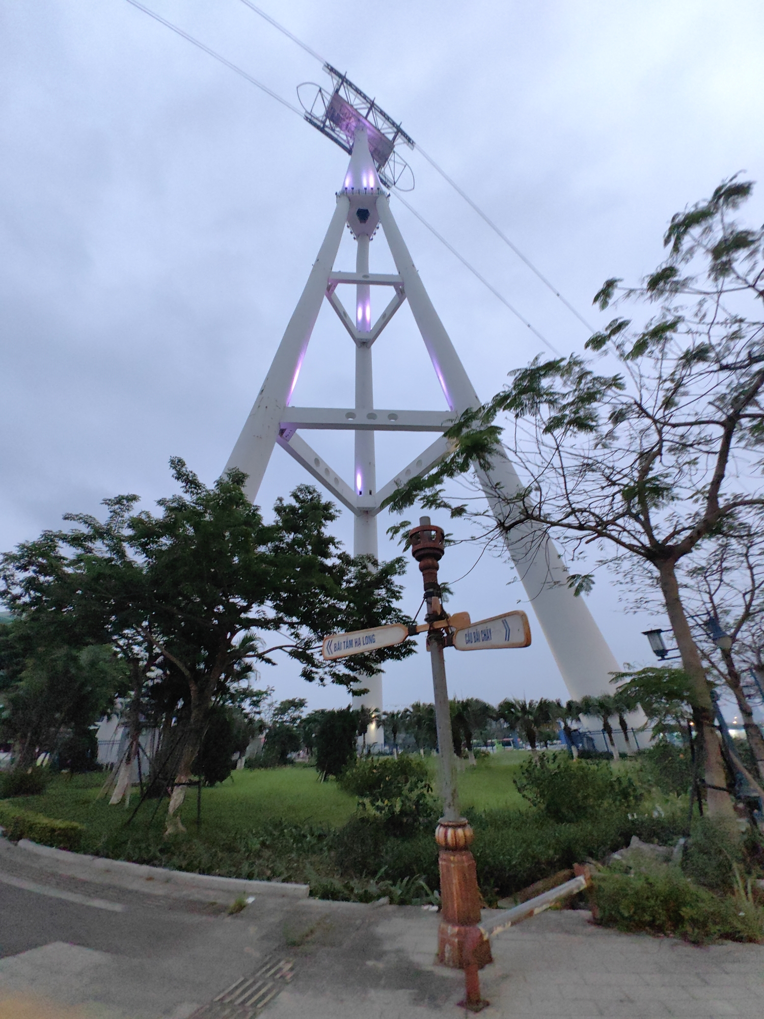 The image displays an outdoor scene at dusk or early evening, characterized by an overcast, light gray sky. The prominent subject is a large, modern cable car pylon, triangular in shape and painted white, which dominates the upper half of the frame. This pylon features internal lighting that emits a soft purple glow from several points along its structure. Cable lines extend diagonally upwards from the pylon, suggesting its function as part of a cable car system.
In the foreground, to the left, a vibrant green tree with dense foliage stands, while another tree with sparser leaves and more visible branches is positioned to the right. A rusty, ornate street signpost is visible in the middle-right foreground. It holds two yellow directional signs: one pointing left with text "BÃI TẮM HẠ LONG" (Ha Long Beach), and another pointing right that reads "CẦU BÃI CHÁY" (Bai Chay Bridge). These signs firmly place the location in Bai Chay Ward, Vietnam.
The lower part of the image features a paved sidewalk with a textured strip, curving from the left towards the right. Adjacent to the sidewalk is a neatly kept grassy area interspersed with various bushes and smaller trees, leading towards the base of the towering pylon. In the background, more distant vegetation and faint lights can be seen. No people or specific activities are depicted, giving the scene a serene and somewhat tranquil ambiance, highlighting the architectural structure against the fading daylight.
