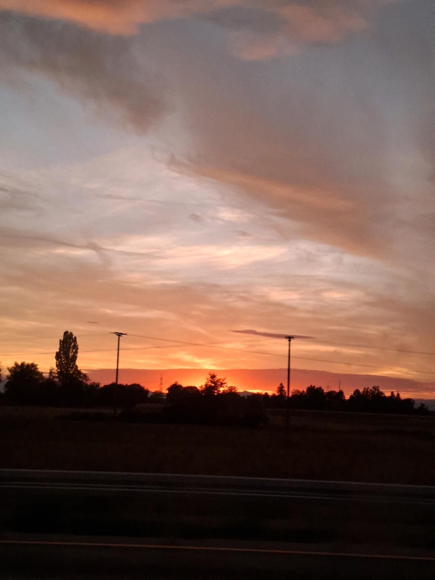 This is a photograph of a sunset over the city of Herbolzheim, Germany. The sky is filled with dramatic clouds, painted in hues of orange, pink, and purple. The sun is low on the horizon, casting a warm glow over the landscape. In the foreground, there is a dark silhouette of trees and a road. A tall, slender tree stands out on the left side of the image, with a lamppost beside it. Further in the distance, silhouetted against the bright sky, is a line of trees and what appears to be a distant tower or industrial structure. The overall impression is one of a serene and colorful end to the day.