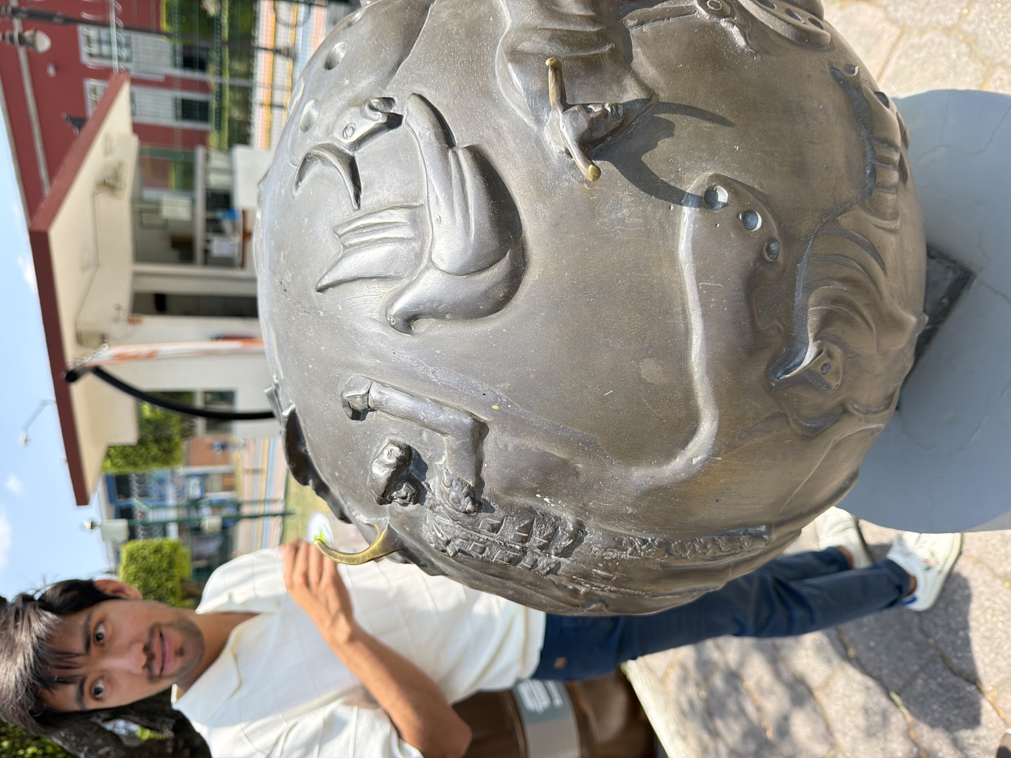 A close-up shot shows a man standing beside a metal globe sculpture. The globe is mounted on a gray base and features raised relief images of ships, animals, and people. The man is wearing a white shirt and blue jeans. He is leaning towards the globe. The location appears to be an outdoor area in San Andrés Cholula, Mexico, indicated by the distinctive building in the background and the architectural style of the globe. The scene suggests a tourist interacting with a local landmark.