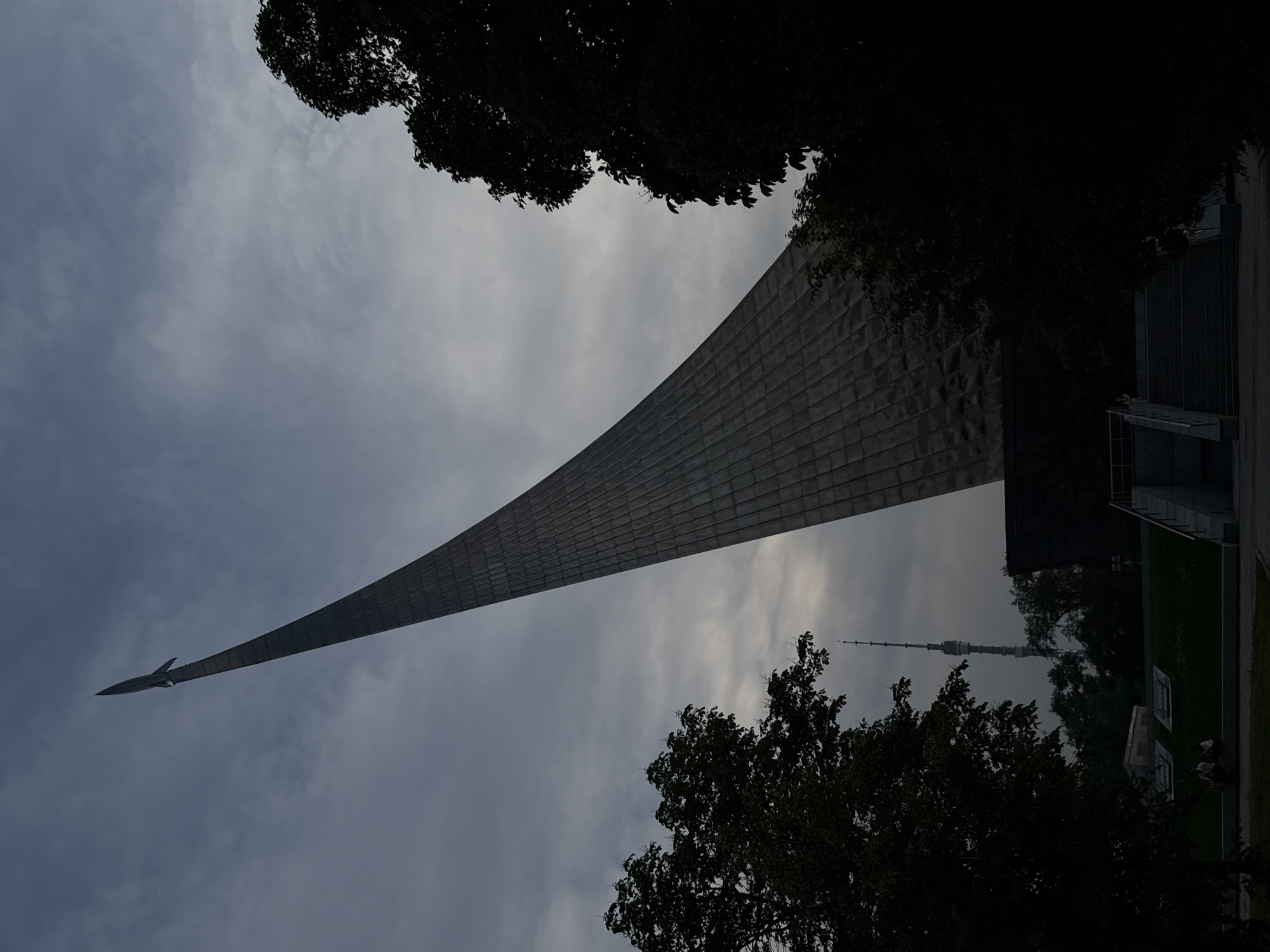 The image depicts the Monument to the Conquerors of Space in Moscow, Russia, under a cloudy sky. The tall, conical monument, designed to resemble a rocket ascending into space, dominates the frame. A small rocket sculpture is visible at its apex. In the background, the Ostankino Tower, another prominent Moscow landmark, can be seen. Dark silhouettes of trees frame the monument on either side, with a portion of the monument's base featuring stairs and a few distant figures seated on a grassy slope. The overall lighting suggests it is either early morning or late evening.
