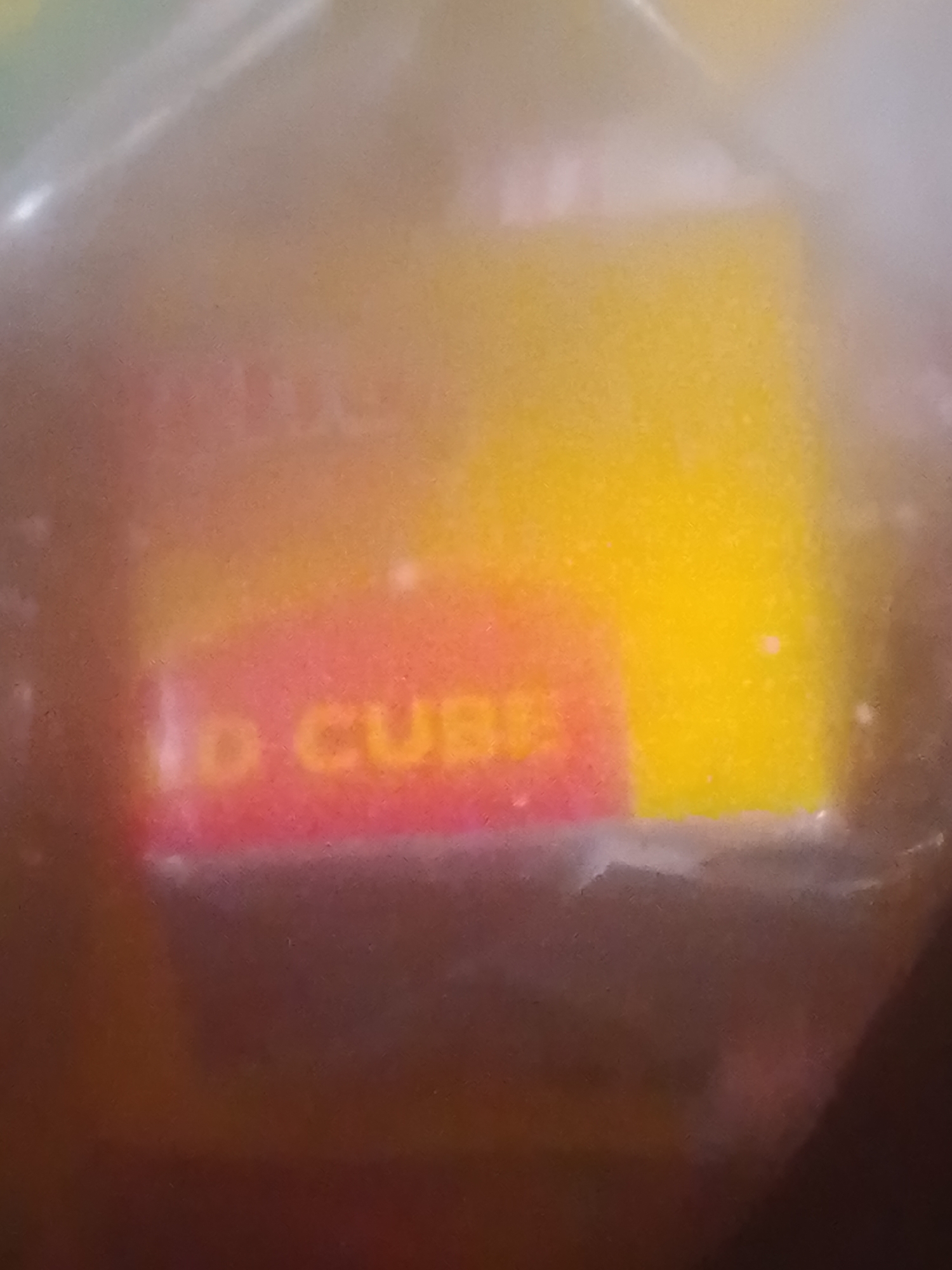 The image shows a close-up, blurry view of what appears to be packaging with text. The text "D CUBE" is visible in orange lettering on a pink and red background. To the right of this, there is a large area of yellow. The overall lighting is bright and diffused, suggesting the item is under direct light and possibly encased in plastic. There is no visible indication of the city of Jalingo, Nigeria, or any specific environmental context. The image is abstract and lacks discernible features of a location or people.
