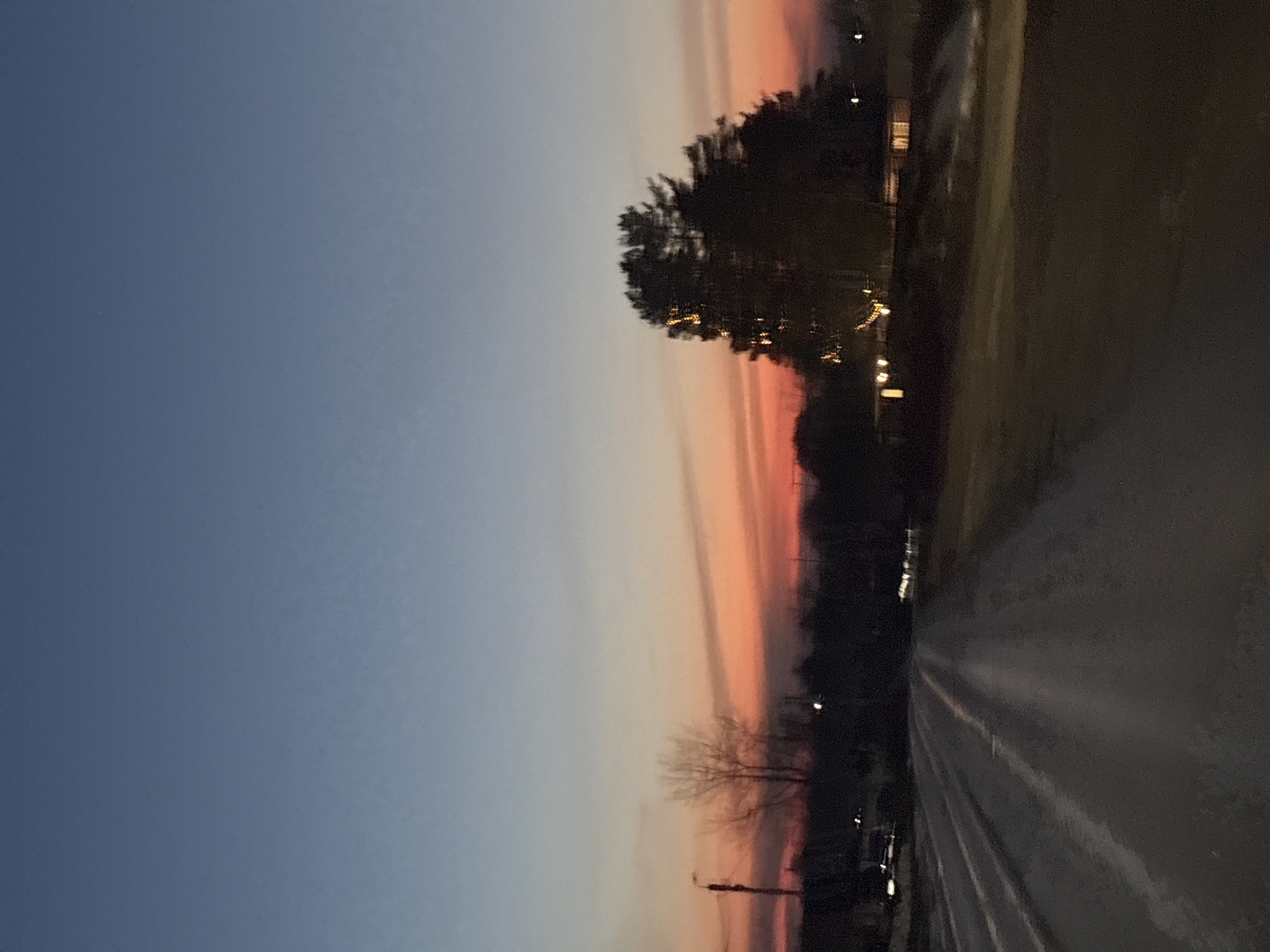 The image depicts a street scene at dusk in Chaikovsky, Russia. The sky transitions from dark blue in the upper left to orange and pink near the horizon. Silhouetted trees and buildings line the side of the road. Streetlights illuminate the road surface, which appears wet. The road leads into the distance, where more lights are visible.