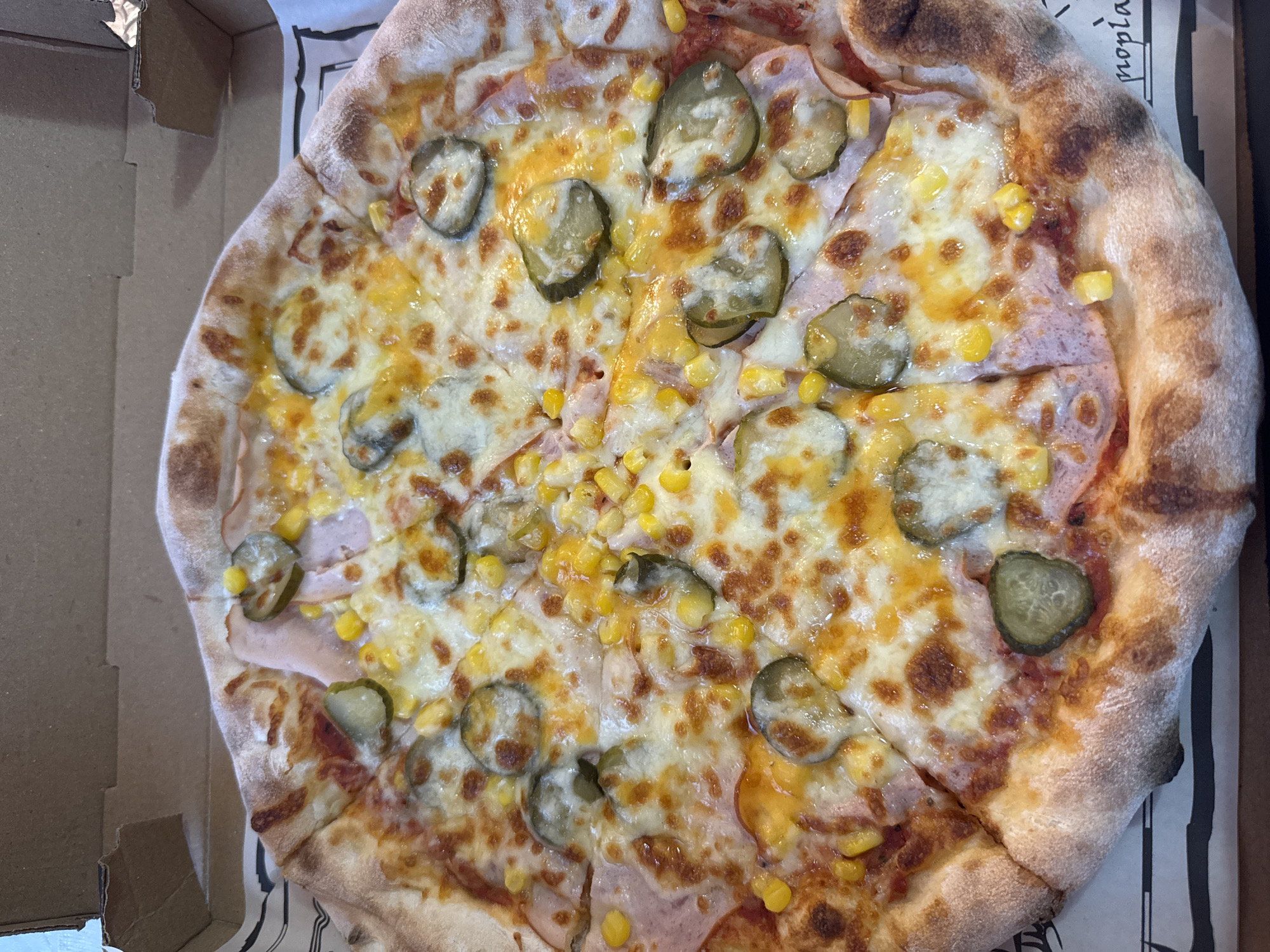 A close-up, overhead shot captures a whole pizza, neatly sliced into eight portions, presented within an open brown cardboard pizza box. The pizza features a well-baked, golden-brown crust with varied textures and some darker, slightly charred spots. The surface is covered with melted, bubbly cheese, displaying a mix of golden-yellow and lighter areas. Distributed across the cheese are numerous circular slices of green pickles (gherkins), scattered bright yellow sweet corn kernels, and visible beneath the cheese, slices of light pink ham. The pizza rests on a white paper liner printed with black graphic patterns and partial text, notably "propia" in a decorative font at the lower right. The surrounding interior of the pizza box is unadorned brown cardboard. No individuals are present in the frame. The scene is brightly lit, focusing entirely on the pizza. Based on provided context, this scene is depicted in the city of Burgas, Bulgaria, though no visual identifiers confirm this specific location within the image itself.