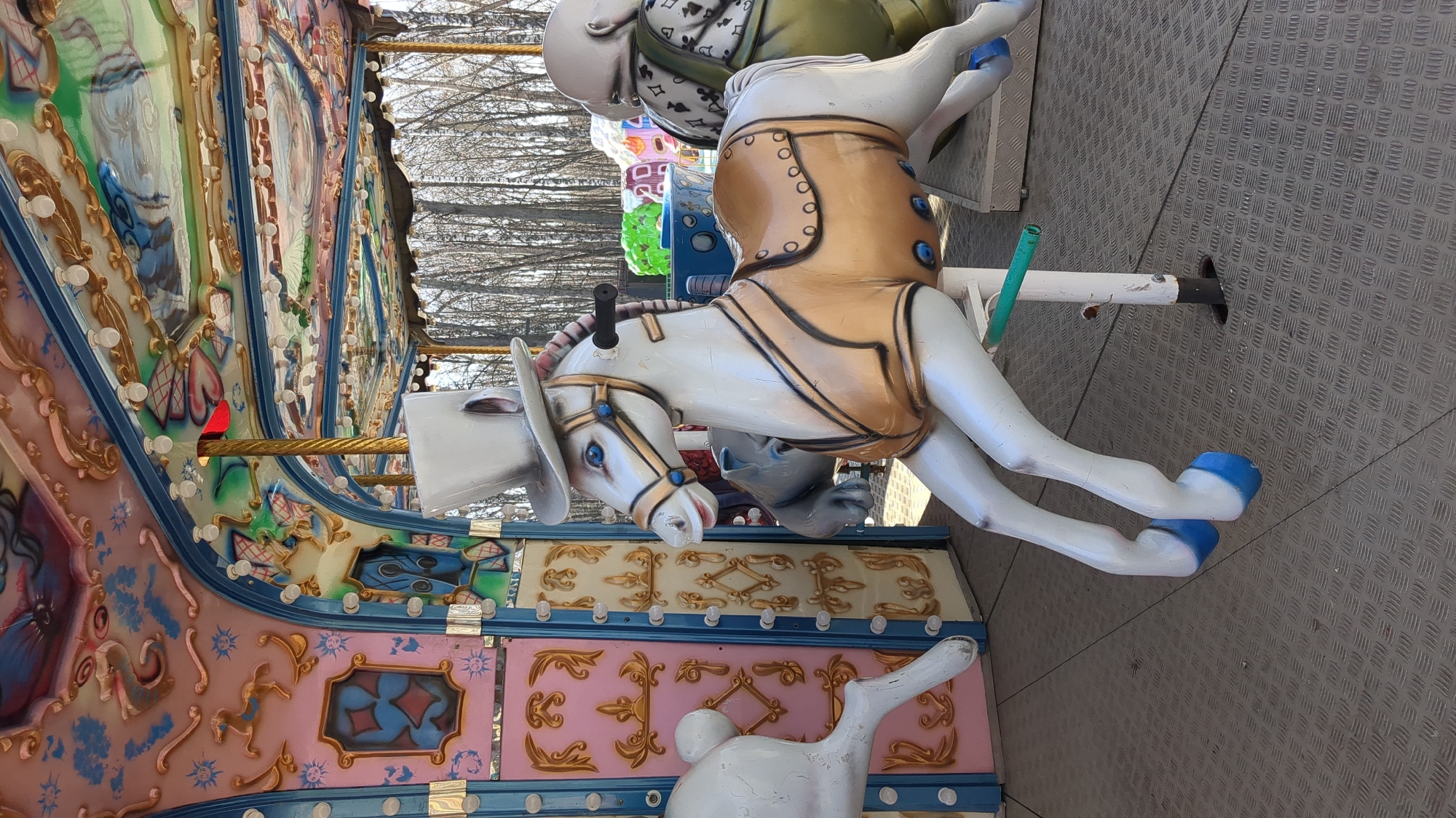 This image captures a close-up view of a stationary carousel, likely located in an outdoor amusement area in Ulyanovsk, Russia. The scene is brightly lit by natural daylight, with bare trees visible in the background, suggesting a cooler season such as late autumn, winter, or early spring.
The main subject is a white carousel horse in the foreground, elaborately decorated with a gold-colored saddle or vest featuring blue buttons, a bridle with blue accents, and a white top hat. Its hooves are painted bright blue. Behind this horse, another carousel figure, possibly a white rabbit, is partially visible, dressed in a green garment adorned with black playing card suit patterns (hearts, spades, diamonds, clubs).
The carousel itself is richly decorated with ornate panels featuring intricate designs in various colors including gold, blue, pink, and red. These panels display illustrative elements such as small horses, hearts, and geometric patterns, with small, clear lights embedded within the trim. The carousel platform is made of a grey, textured metal with a diamond plate pattern. Support poles for the carousel figures are also visible. There are no people visible, indicating the carousel is not currently in operation. No legible text is visible in the image.