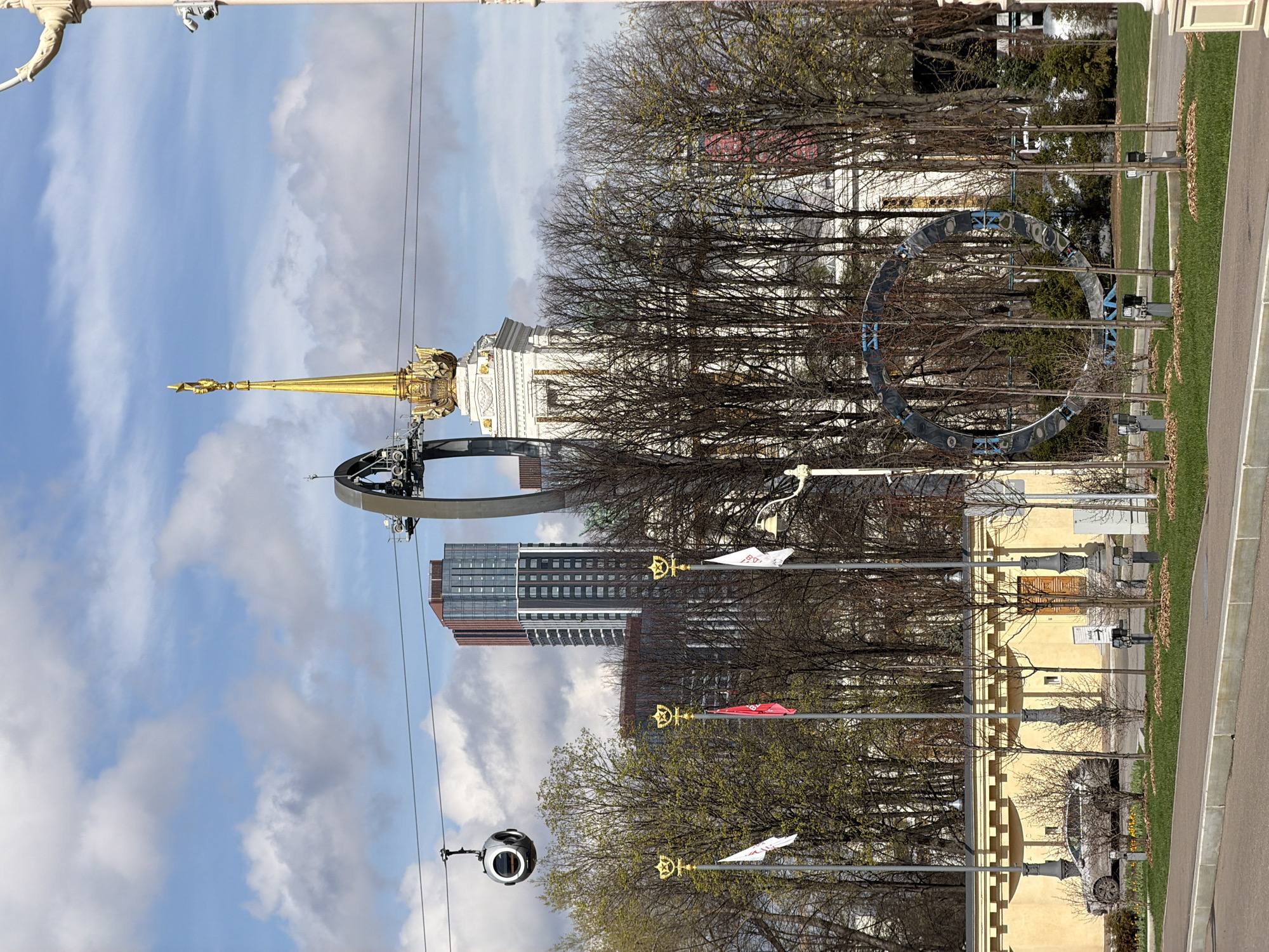 The image captures a dynamic urban landscape, likely the VDNKh (Exhibition of Achievements of National Economy) in Moscow, under a partly cloudy blue sky. In the foreground, a dark, spherical cable car gondola hangs from its wire, suggesting an active transport system.
The scene features a striking blend of architectural styles. A magnificent golden spire, characteristic of Stalinist-era architecture, rises prominently from a large, ornate white building. To its right, a monumental dark metal circular structure, possibly an art installation or part of a ride, adds a contemporary sculptural element. Behind these, a tall, modern high-rise building with a dark facade and numerous windows stands out against the horizon.
Rows of trees, mostly bare but with hints of early spring foliage, line the pathways, separating the various structures. Below them, a paved road or path runs alongside strips of neatly maintained green grass. Several streetlights, topped with golden stars, display flags. One white flag clearly shows "ВДНХ 80" in blue text, while a red flag partially visible behind it appears to commemorate "1939-2019," indicating the 80th anniversary of VDNKh. Further down the path, a yellow, classically styled building with a brown wooden door is visible, with a silver car parked nearby.
The time of day is clearly daytime, with bright sunlight illuminating the scene. No specific human activity or event is distinctly captured beyond the operational cable car and the commemorative flags. The overall impression is one of an expansive park or exhibition complex, showcasing both historical grandeur and modern development.