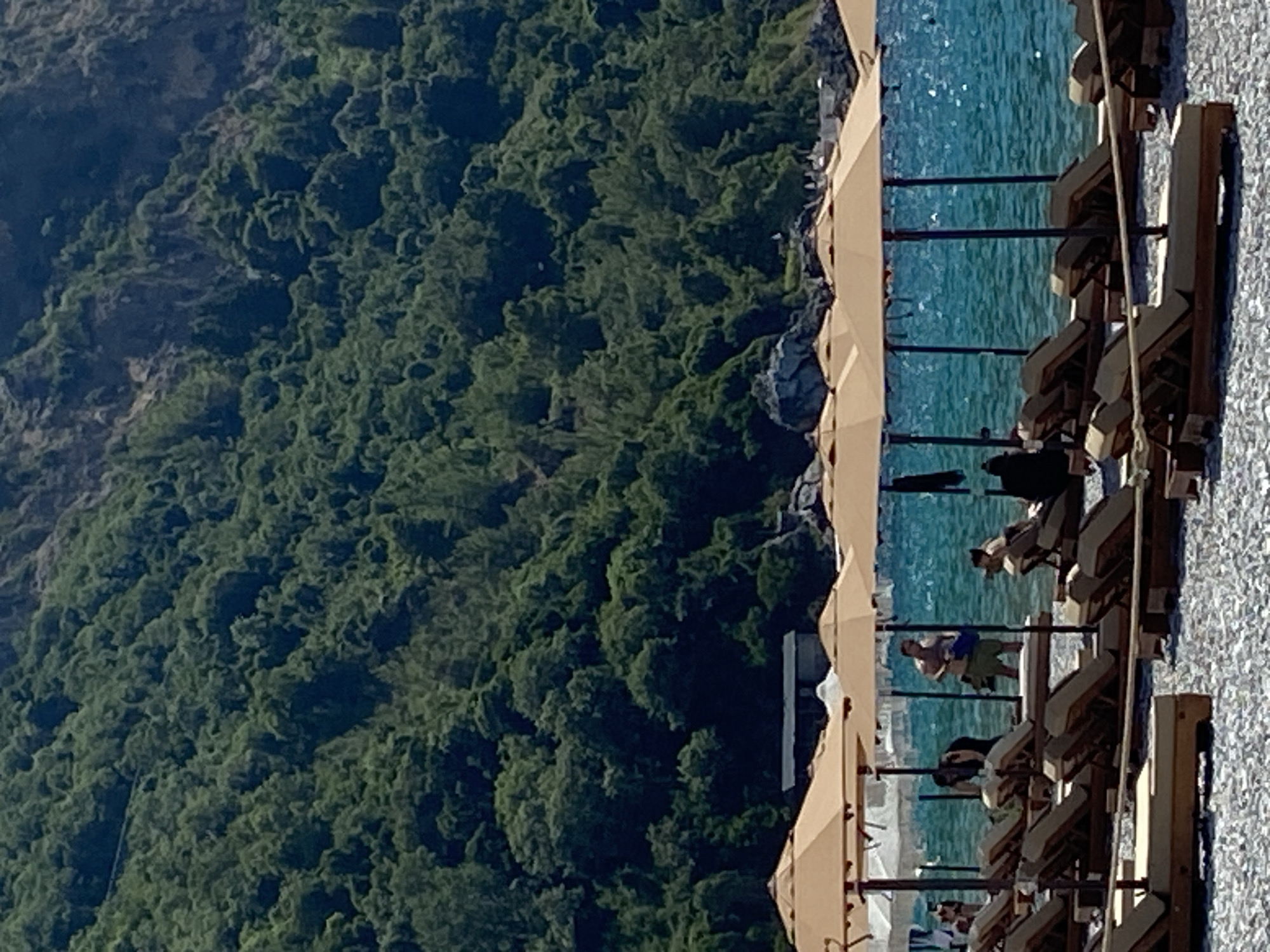 This image depicts a coastal scene in Prijevor, Montenegro, featuring a beach with rows of unoccupied lounge chairs and umbrellas. The beach is covered in pebbles. In the background, a lush, green, forested mountainside rises steeply, with visible rocky outcrops. The turquoise sea stretches across the midground, and several people are visible near the water's edge, some standing and others partially obscured by umbrellas. One person, a male in shorts, is standing and holding a blue bag. Another person, a female, is partially visible to the left of him. A few more individuals are seen seated or standing further back, near the water. A small, dark-colored structure is situated on the hillside behind the beach umbrellas.