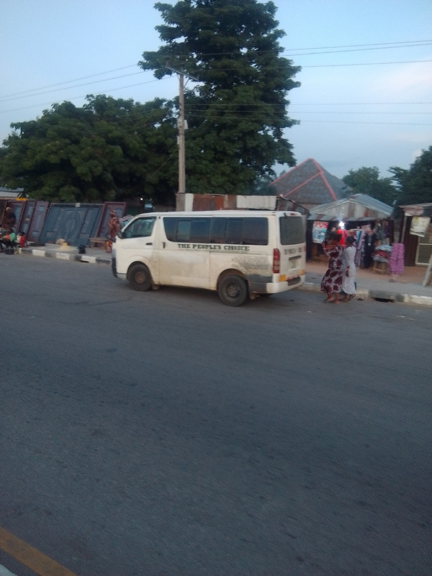 A white van with "THE PEOPLE'S CHOICE" written on the side is parked on the side of a road in Gulum, Nigeria. The van is dirty and appears to be well-used. Two women are standing near the van, one wearing a patterned dress and the other a white dress. In the background, there are several stalls or shops with goods displayed, suggesting a market area. Trees and utility poles are also visible. The sky is overcast, indicating it might be late in the day or during a period of bad weather.