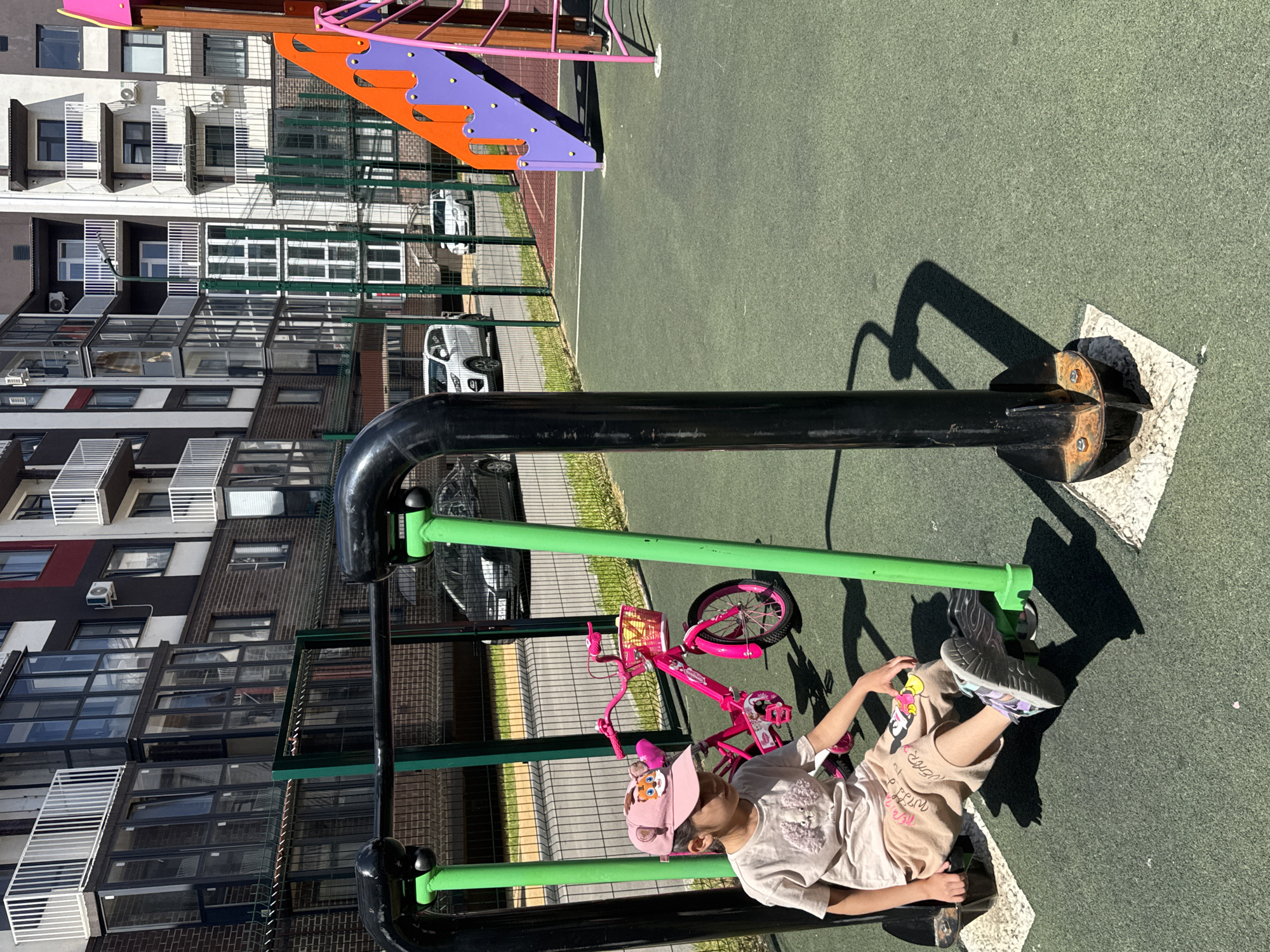 A young girl is sitting on a playground exercise machine in a city park in Vyazniki, Russia. She is wearing a light-colored t-shirt, light brown shorts with cartoon characters, and a pink baseball cap with a tiger design. Her feet are on the machine's footrests, and her hands are resting on the handles. To her right, a pink children's bicycle with a basket is parked. Behind the playground equipment, a fence separates the park from a residential building with multiple balconies and windows. Several cars are parked in front of the building. The ground is covered with green rubberized material, and the sky is clear and blue.