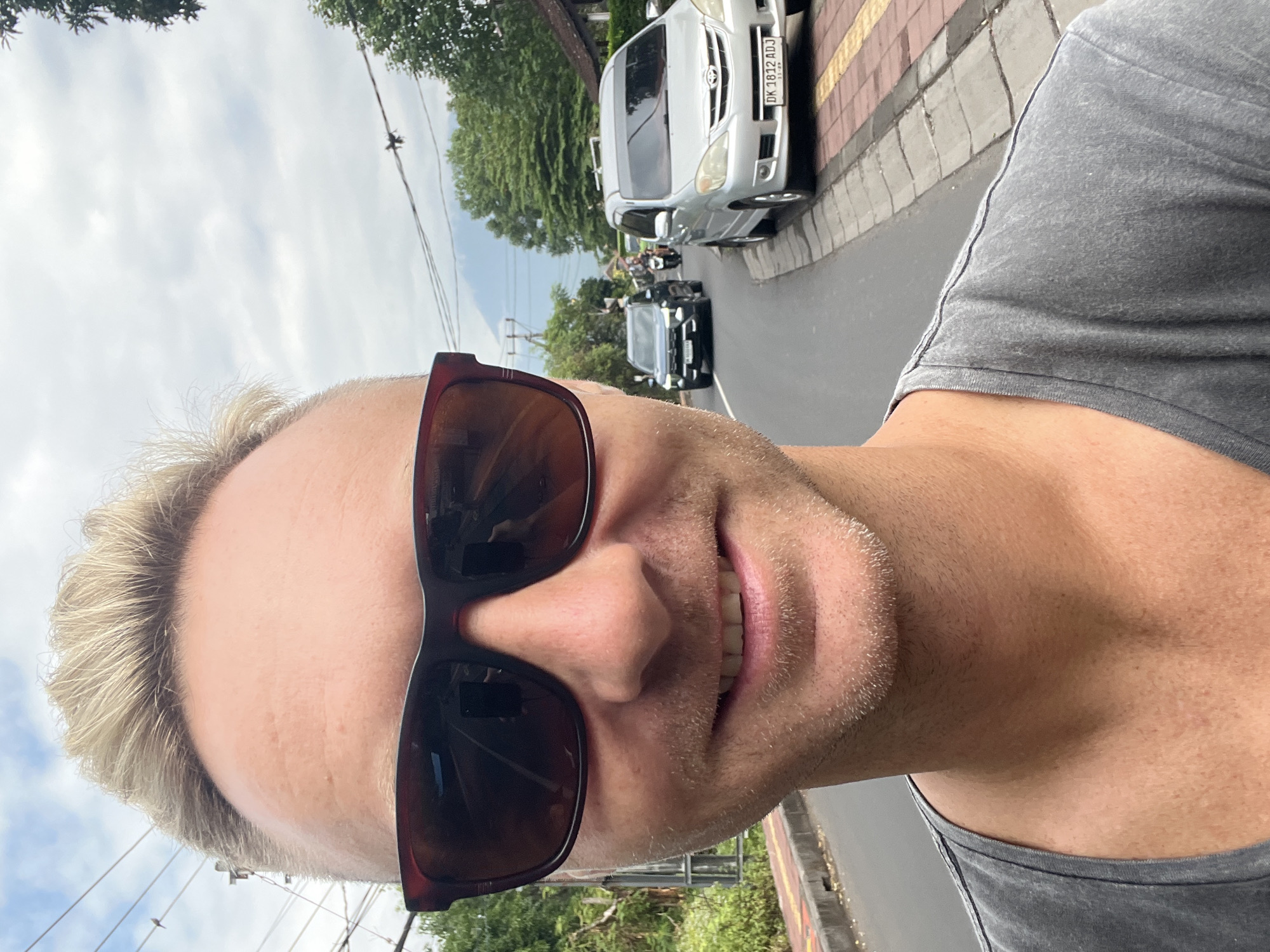 A man with blond hair and sunglasses is taking a selfie in front of a street scene in Abang, Indonesia. He is wearing a gray t-shirt. In the background, there is a road with a silver car on the right and a dark SUV on the left. There are also motorcycles and trees visible. The sky is cloudy.
