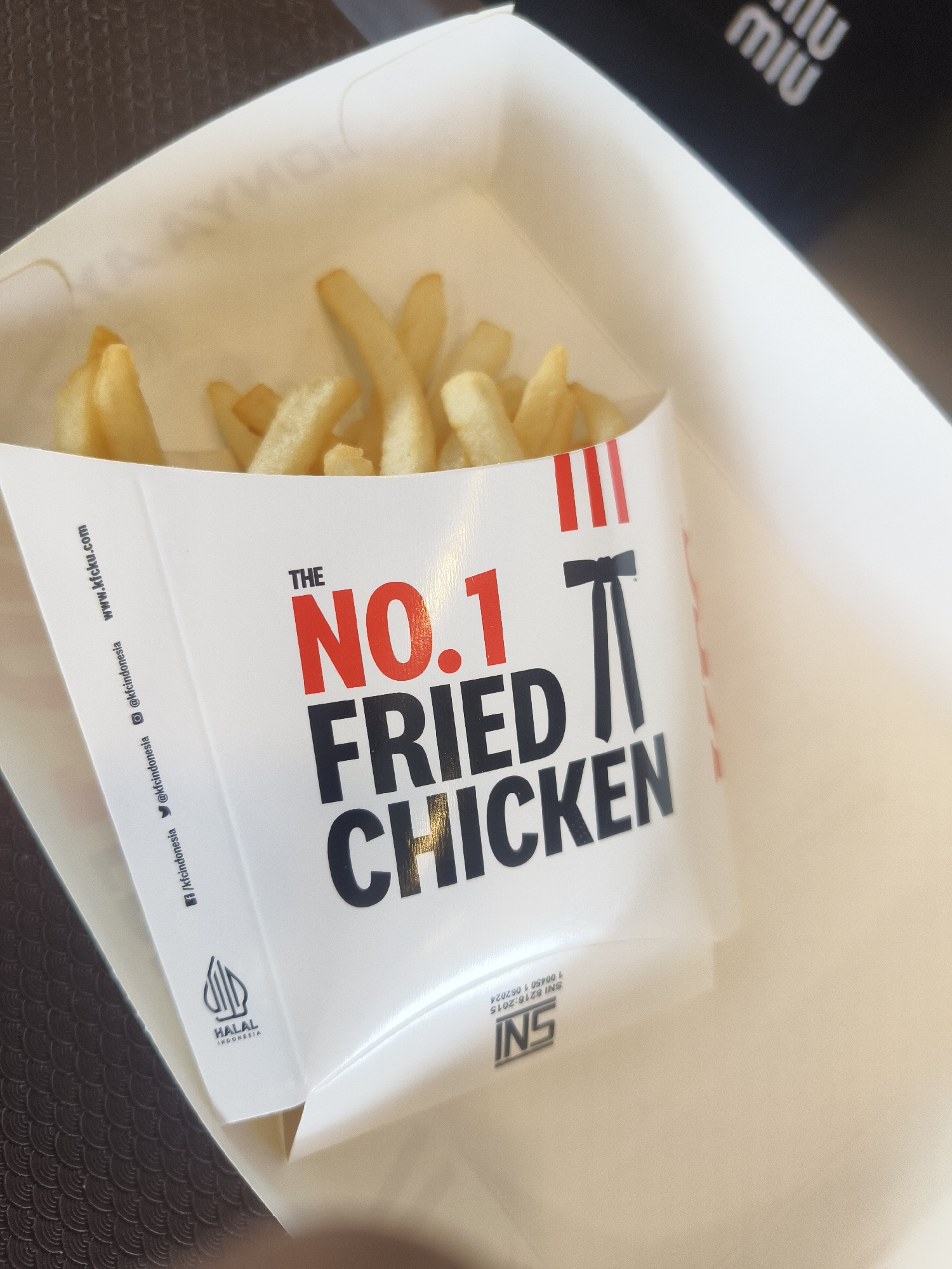 The image shows a close-up of a white paper box filled with french fries. The box has text and logos printed on it. The text "THE NO. 1 FRIED CHICKEN" is prominently displayed in red and black lettering. There are also social media handles such as "@kfcindonesia" and a website address "www.kfc.com" printed on the side of the box. A "HALAL INDONESIA" logo is visible on the lower left side of the box. The box is placed on a dark, textured surface that appears to be a table. In the background, a blurry black object with white logos resembling "miu miu" is visible. The overall lighting suggests an indoor setting. There are no people or animals visible in the image. The image is a product shot of a food item.