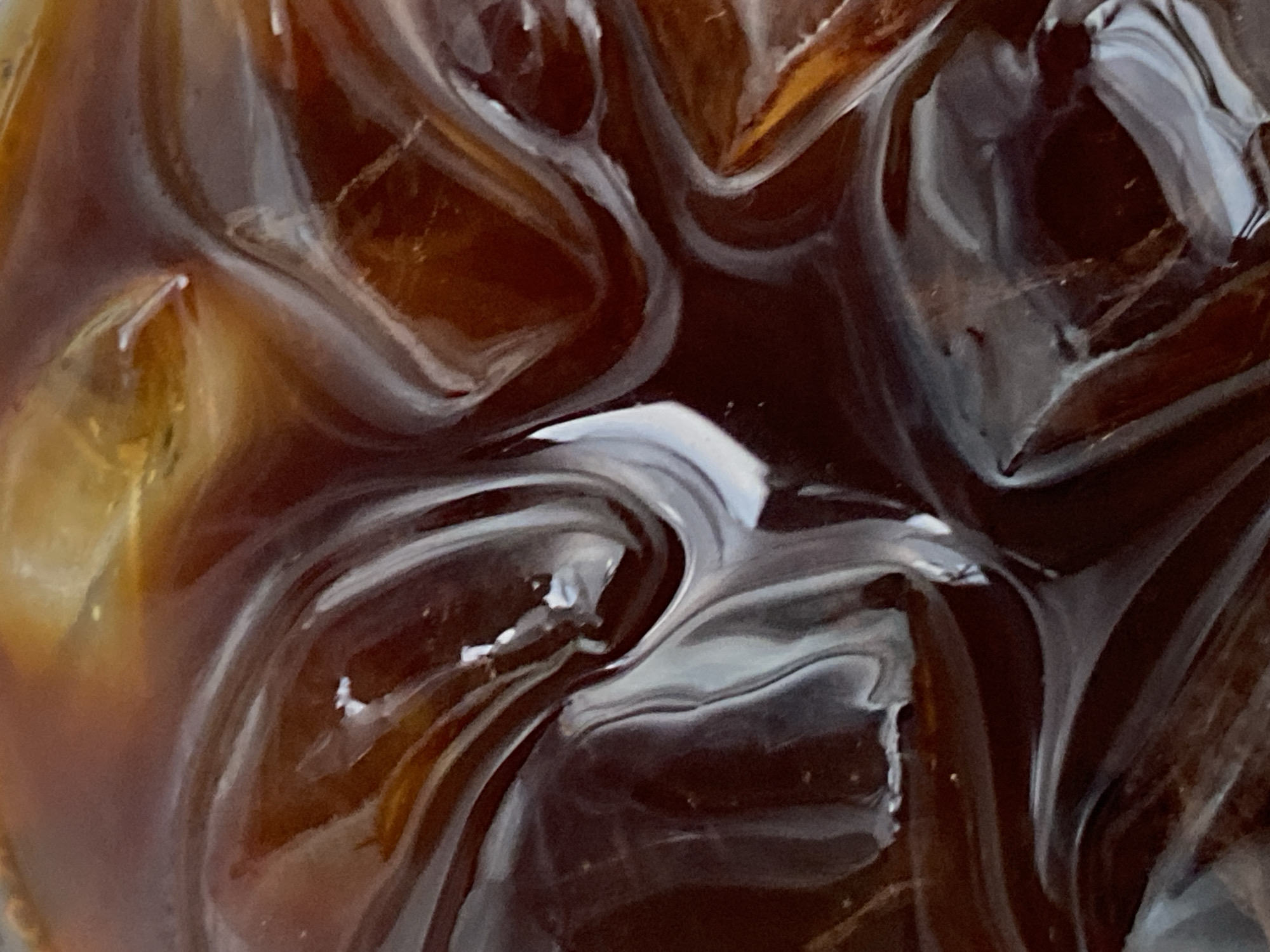 This is an extreme close-up, macro photograph of iced coffee. The image is dominated by the dark brown liquid and translucent ice cubes. Swirls of lighter brown and white are visible, suggesting the mixing of coffee and cream or milk, or possibly light reflecting off the ice and liquid. The texture is smooth and glossy, with sharp lines and rounded shapes formed by the ice. There are no discernible people, objects, or environmental features outside of the beverage itself. The location in Mengwi, Indonesia, is irrelevant to the visual content of the image.