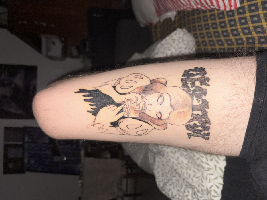 The image displays a human leg, located in Tel-Aviv, Israel, extending diagonally from the lower left to the upper right of the frame. The leg presents fair skin with visible hair. A multi-component tattoo is prominently featured on the leg. The central element of the tattoo is an inverted, cartoon-style female figure with closed eyes, reddish-brown hair, and large, rounded, ear-like structures. Her face and hair are rendered in color, while other elements are delineated with black outlines. Above this inverted figure (relative to the tattoo's orientation), two hands are depicted reaching downward, appearing to manipulate or break a thin, rectangular object. Further above, a solid black, jagged silhouette suggesting a cityscape or mountains is present. Below the inverted figure, the word "MALFEAD" is tattooed in a dark, stylized, and somewhat distressed font. The lower portion of the leg is covered by dark fabric, consistent with shorts or underwear. The background reveals a dimly lit indoor environment, likely a bedroom. On the left, light-colored bedding with a subtle pattern is visible, while on the right, darker, geometrically patterned bedding is present, alongside a partially visible red and white striped object. Indistinct furniture and a framed artwork are discernible in the blurred far background.
