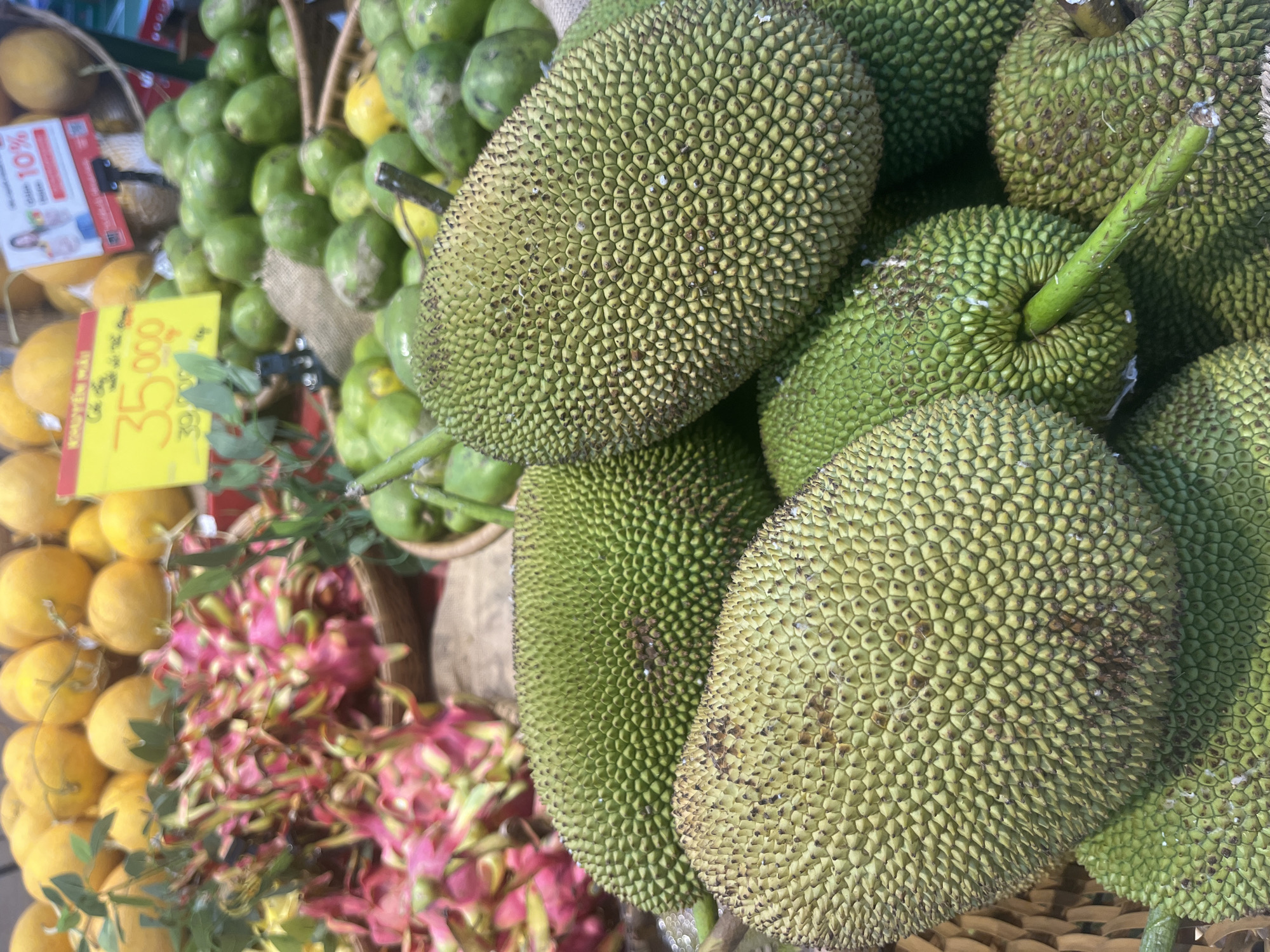 The image shows a close-up view of a pile of large, green jackfruits. The jackfruits have a spiky exterior with a pattern of hexagonal scales. The fruits are stacked on top of each other in a woven basket. In the background, there are other fruits visible, including yellow citrus fruits and pink dragon fruits. A yellow price tag is also visible in the background, with Vietnamese text and the number "35.000" and "39.000" printed on it. The lighting suggests an indoor setting, possibly a market.