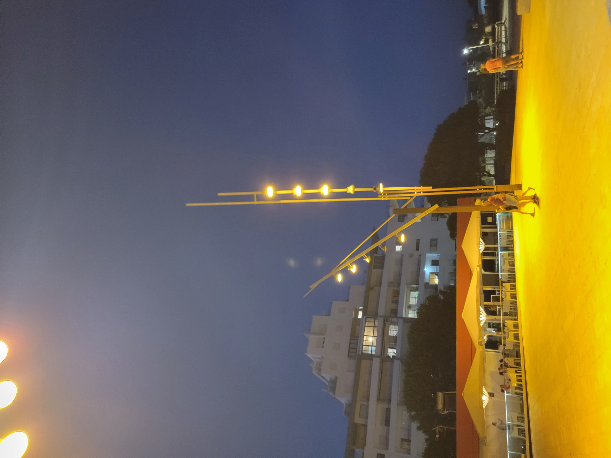 This is a nighttime vertical, full-shot photograph taken in Quarteira, Portugal, looking towards a modern, multi-story white building with many windows. The sky is a deep blue, transitioning to a lighter hue near the top where there are several out-of-focus yellow lights visible. In the foreground, a tall, modern yellow lamppost with multiple lights stands on the right, angled slightly towards the left of the frame. To the left of the lamppost, and partially obscured by it, is a white building with orange awnings and outdoor seating. Several people are seated at tables under the awnings. In the middle ground, two figures are walking on a bright yellow, illuminated paved area. In the background, behind the lamppost and to the right, are more illuminated paved areas with benches and some distant lights. The overall scene depicts a lively outdoor public space at night, likely a promenade or a restaurant area, with the modern architecture of Quarteira in the background.