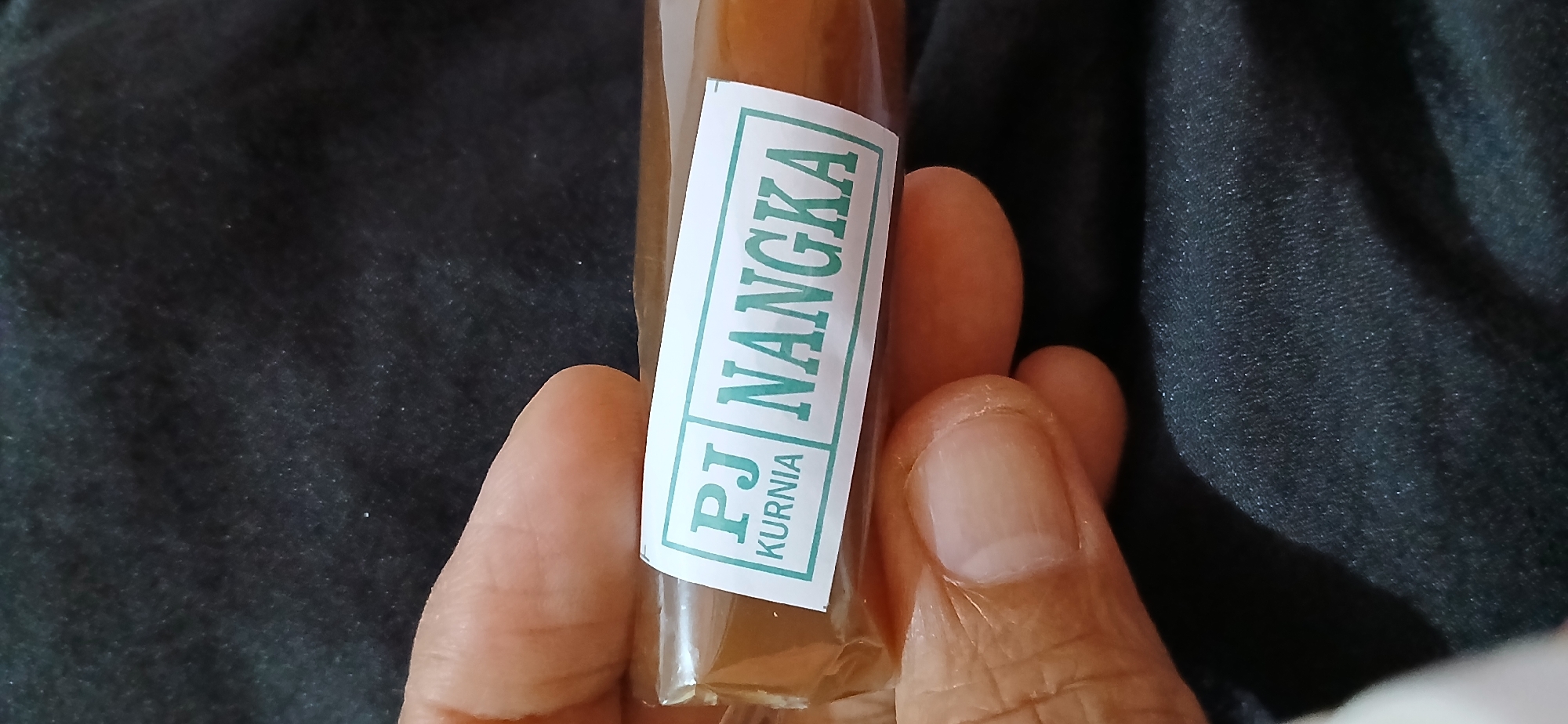 A close-up, eye-level shot captures a hand holding a packaged food item. The item is wrapped in clear plastic and features a white label with green text. The label reads "PJ KURNIA NANGKA". The background is a dark, textured fabric, possibly clothing or upholstery. The image is undated and lacks specific location information within Samarinda, Indonesia.