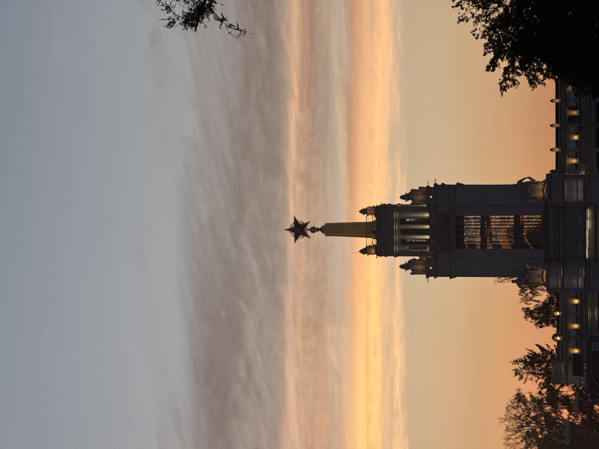 The image depicts the Kotelnicheskaya Embankment Building in Moscow, Russia, at sunset. The sky is striated with orange and gray clouds. The building is illuminated and topped with a spire and star. Silhouetted tree branches frame the building.