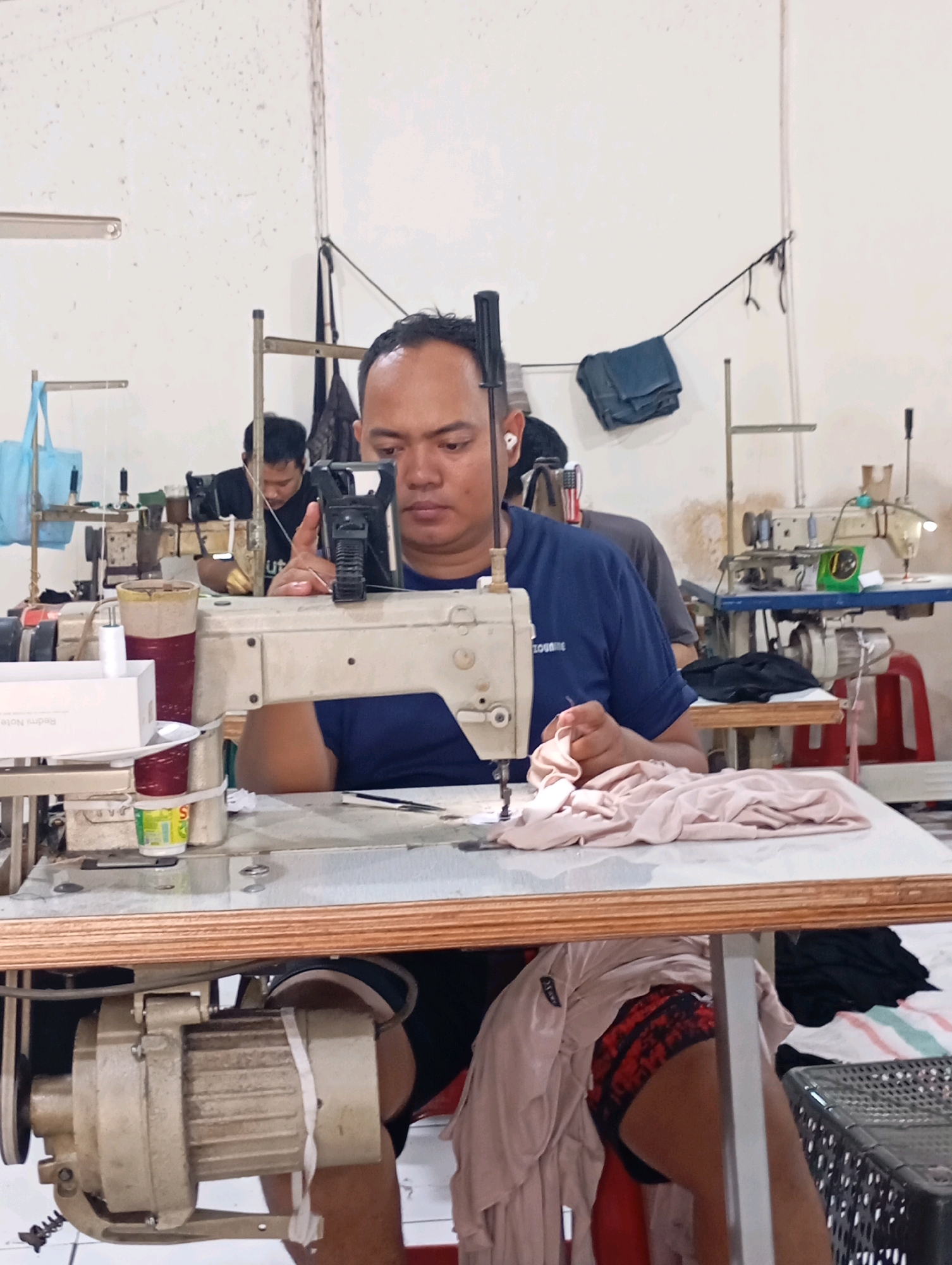 The image depicts an indoor sewing workshop in Tasikmalaya, Indonesia. A man in a blue t-shirt is seated at a sewing machine, actively engaged in sewing. He is wearing earbuds and appears focused on his work, holding a piece of light pink fabric. Several sewing machines are visible in the background, along with other individuals working. The environment suggests a functional, perhaps slightly cluttered, workspace with various sewing supplies and materials scattered around. The overall impression is of a busy production setting.