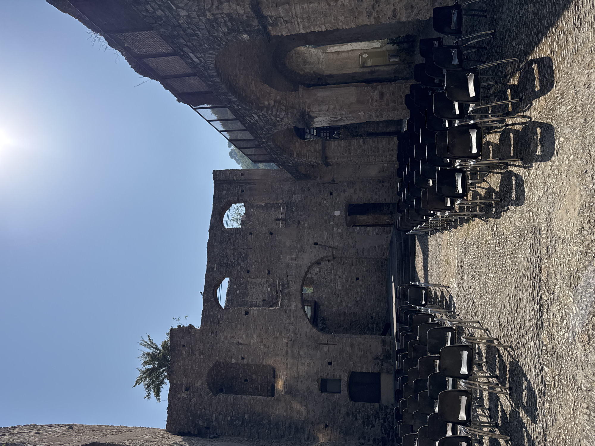 The image shows an outdoor area in Dolceacqua, Italy, featuring rows of empty black plastic chairs arranged on a cobbled ground. Behind the chairs, the remains of ancient stone structures are visible, with arches, openings, and crumbling walls. The ruins suggest a historical or archaeological site, possibly a former amphitheater or performance space, given the seating arrangement. Sunlight casts shadows from the chairs onto the ground, and a bright sun is visible in the upper right portion of the sky. The architecture and natural elements indicate a warm, sunny day in an old European town.