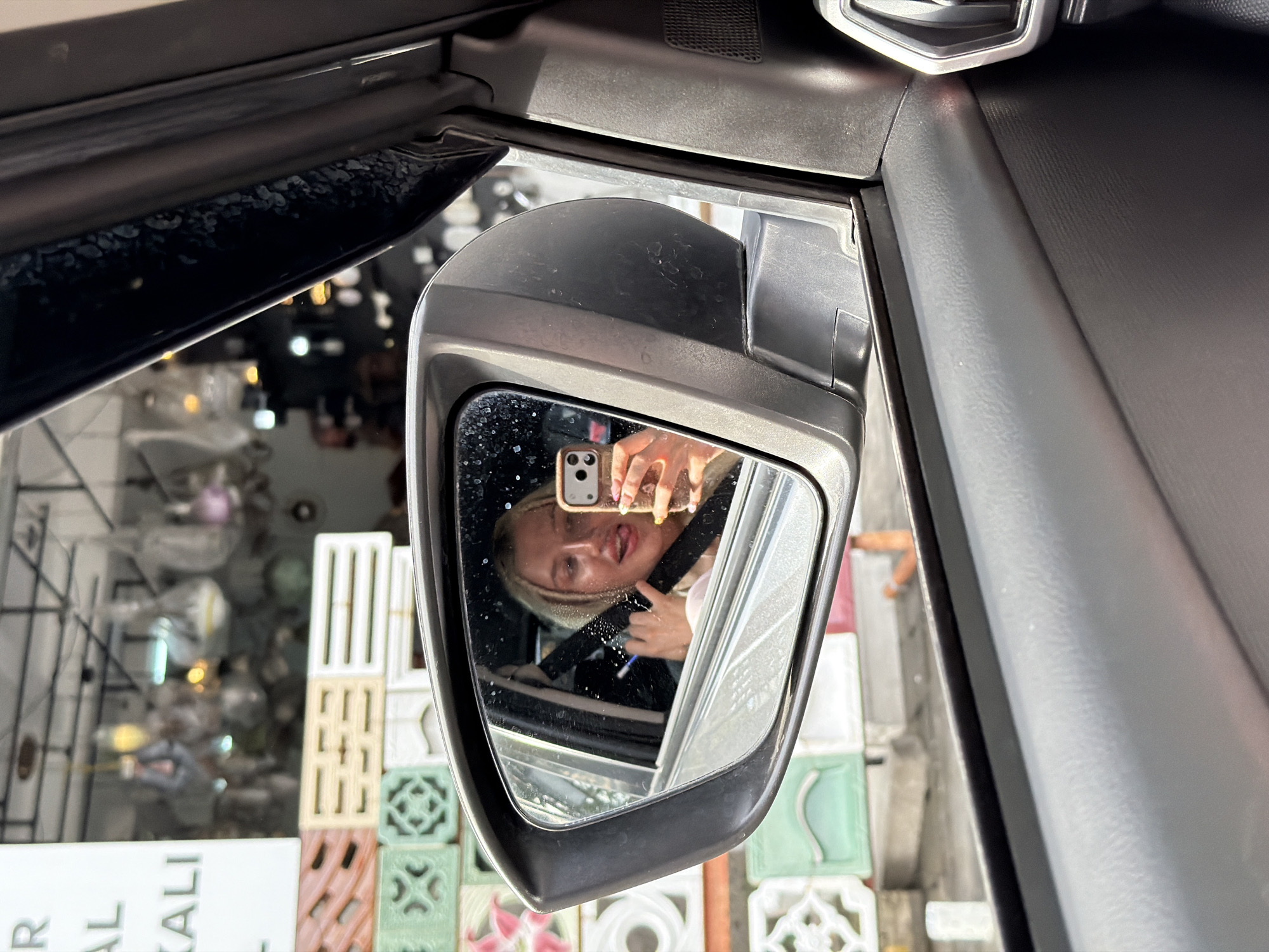A young woman with blonde hair is visible in the side mirror of a car, holding up a smartphone and taking a selfie. She is sticking out her tongue and making a peace sign with her fingers. The car appears to be parked on a street in Mengwi, Indonesia, with a store selling decorative tiles visible in the background. The tiles are stacked in various colors and patterns, and several light fixtures are hanging from the ceiling of the store. The car's interior, including the dashboard and door panel, is also visible in the foreground. The overall scene suggests a candid moment captured during a casual outing in a bustling urban environment.