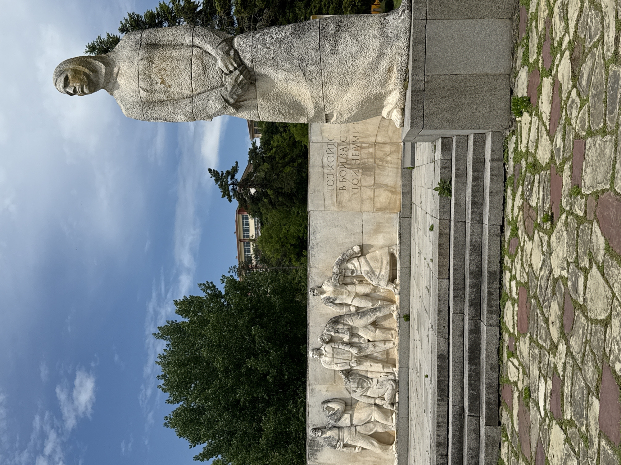 The image shows a stone monument in Strazhitsa, Bulgaria. Atop the monument is a weathered stone sculpture of a woman. Below the statue, a stone plaque bears an inscription. Beneath this plaque is a frieze depicting five figures in stone relief. In the background, a tree line and a light-colored building are visible beneath a blue sky with scattered clouds. A set of stone steps leads up to the monument, and a stone-paved area borders the monument's base. The image has been rotated 90 degrees counterclockwise.