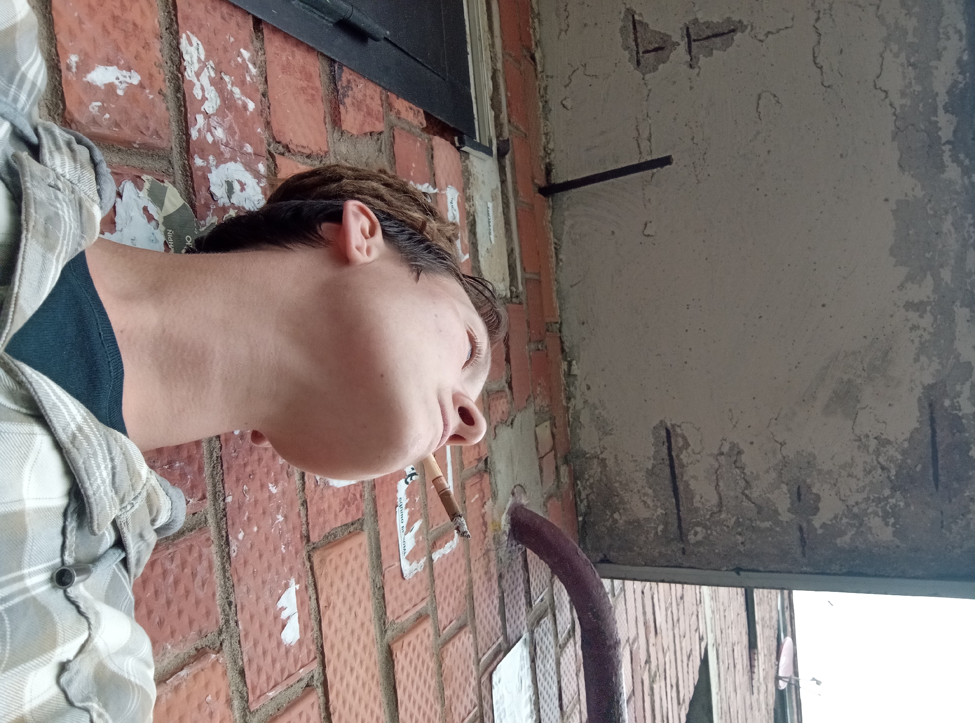 A low-angle medium shot captures a person smoking a cigarette outdoors against a brick wall and a concrete structure. The person's head is tilted back, and their eyes are closed, with smoke exhaling from their mouth. They are wearing a plaid shirt over a dark t-shirt. The background consists of a rough, weathered concrete overhang with exposed rebar, a red brick wall with remnants of posters, and a large, dark metal door on the left. A thick, dark pipe curves into the scene from the right. The overall environment suggests an urban or semi-urban setting. No specific location in Gdov, Russia, can be determined from the visual information.