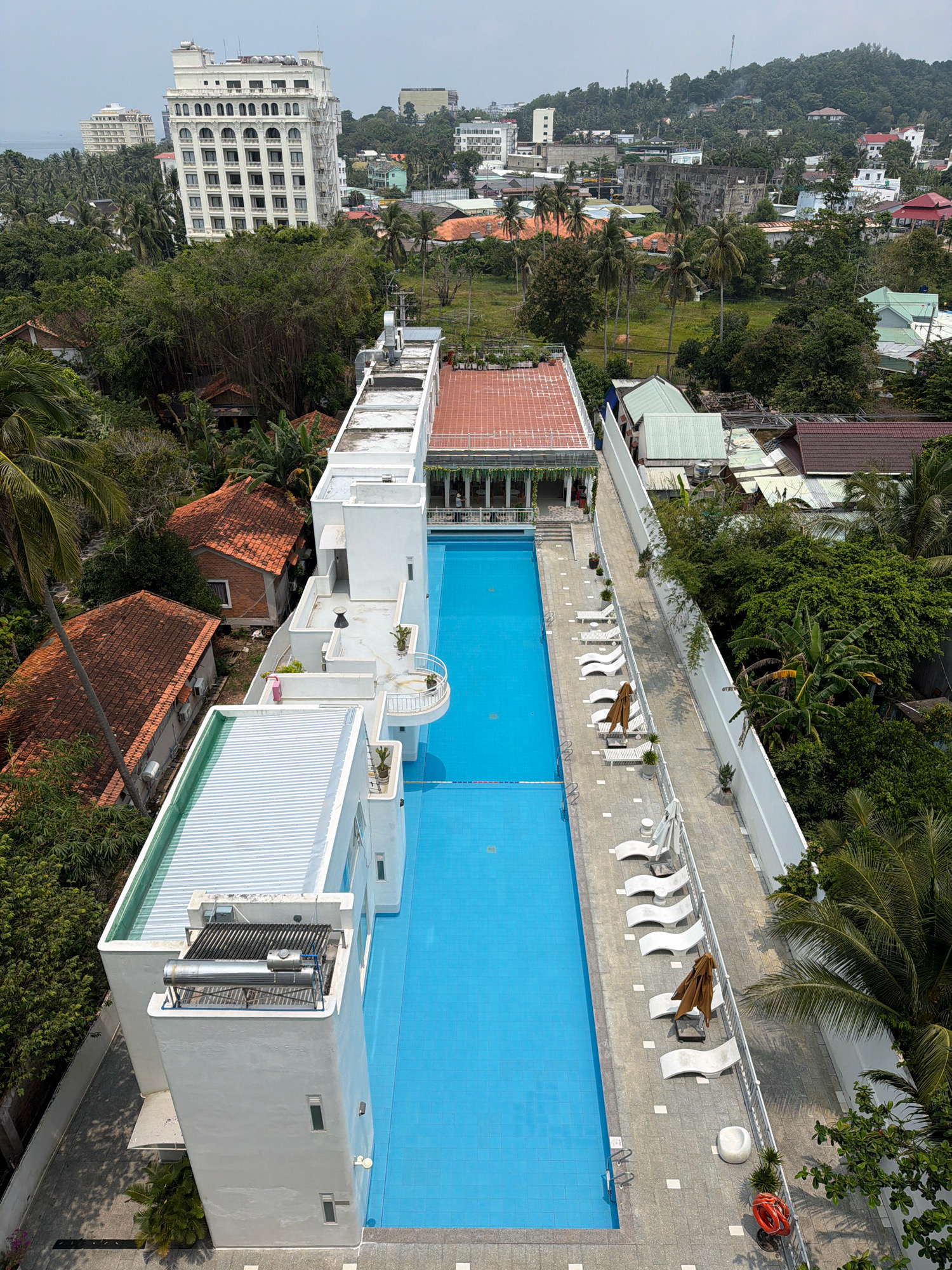 An aerial view shows a resort complex with a long, bright blue swimming pool as the central feature. The pool is surrounded by white lounge chairs, some with open brown umbrellas, arranged neatly along a grey tiled walkway. Adjacent to the pool is a white multi-story building with balconies and many windows. To the left of the pool area are several traditional Vietnamese houses with terracotta tile roofs, nestled amongst lush green tropical trees and palm trees. In the background, a larger, white hotel or apartment building stands out, with other buildings and green hills in the distance, suggesting an urban or semi-urban setting in Phú Quốc, Vietnam. The sky is overcast.