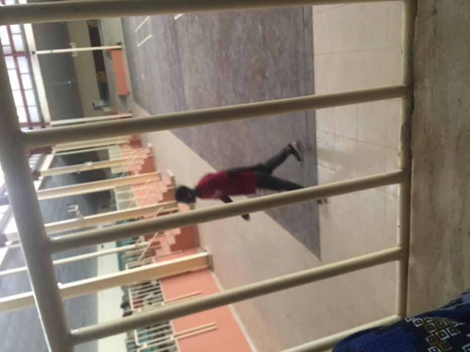 The image depicts an indoor facility, located in Pindiga, Nigeria, viewed through light-colored vertical and horizontal railings from a slightly elevated position. In the foreground, the lower-left corner shows a portion of a person's shoulder and arm, dressed in a blue patterned garment, indicating the photographer's viewpoint.
In the midground, a single male individual is captured in motion, walking from left to right. He is wearing a red short-sleeved shirt, dark trousers, and dark shoes. His figure is somewhat blurred, consistent with movement. He is traversing a light-colored tiled floor, which transitions into a darker, rectangular floor area on the right side of the frame.
The background reveals a large internal space, possibly a gymnasium or auditorium. Along the left and receding towards the rear, multiple tiers of spectator seating or bleachers are visible, characterized by light-colored railings and orange-painted lower walls and steps. Several vertical, light-colored support columns are interspersed throughout this architectural feature. The far background shows a vast open floor area, possibly an indoor court, beneath a high ceiling structure. The scene is evenly lit.
