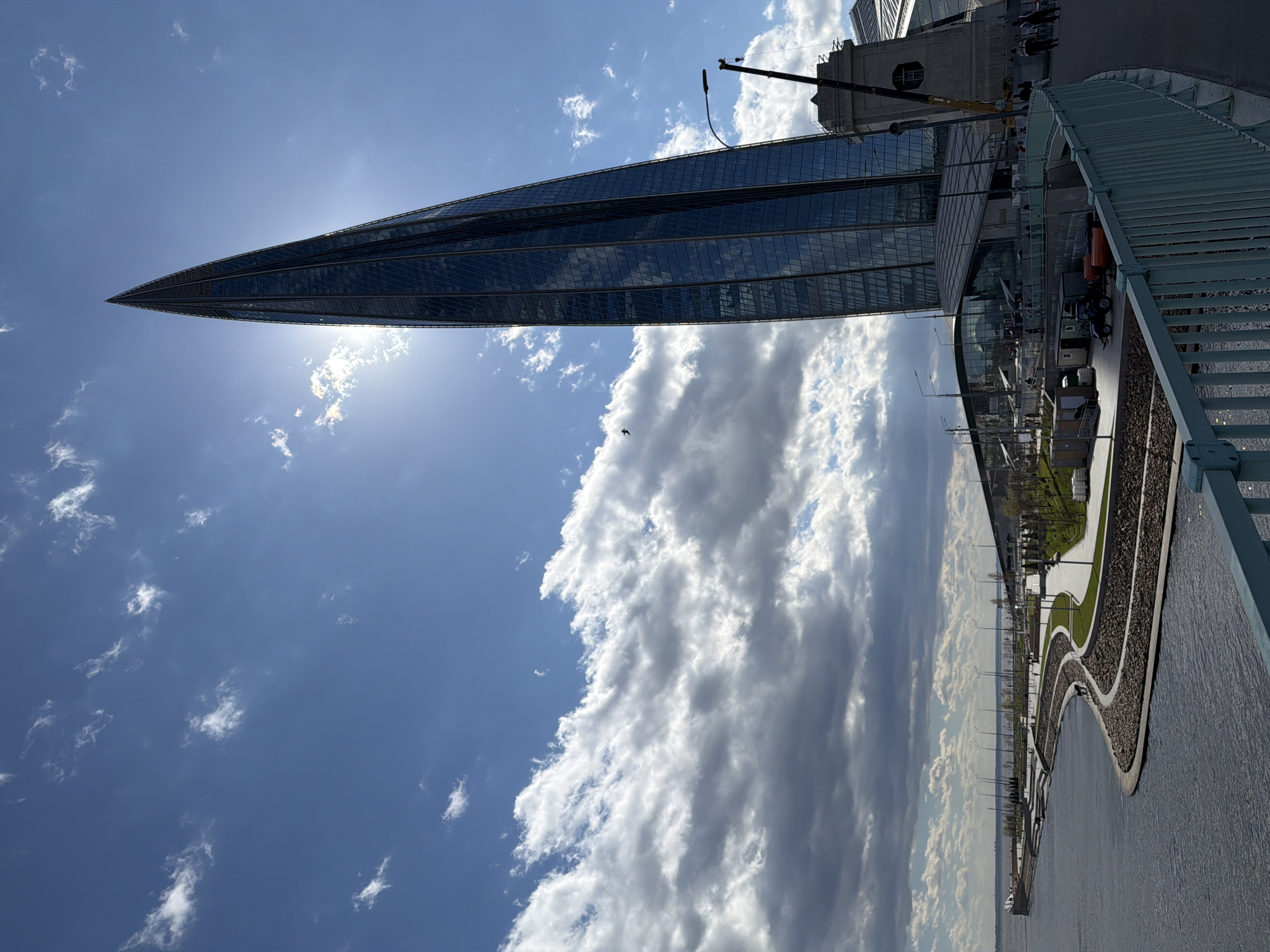 The image depicts the Lakhta Center, a skyscraper in Saint Petersburg, Russia, silhouetted against a bright, partly cloudy sky. The building is a tall, pointed structure with a glass facade reflecting the sunlight. Part of a bridge is visible in the foreground, and a landscaped area, possibly a park, with curved paths is present in the middle ground. There's a body of water in the lower part of the image. A construction crane is visible on one side of the building. A small bird is flying.