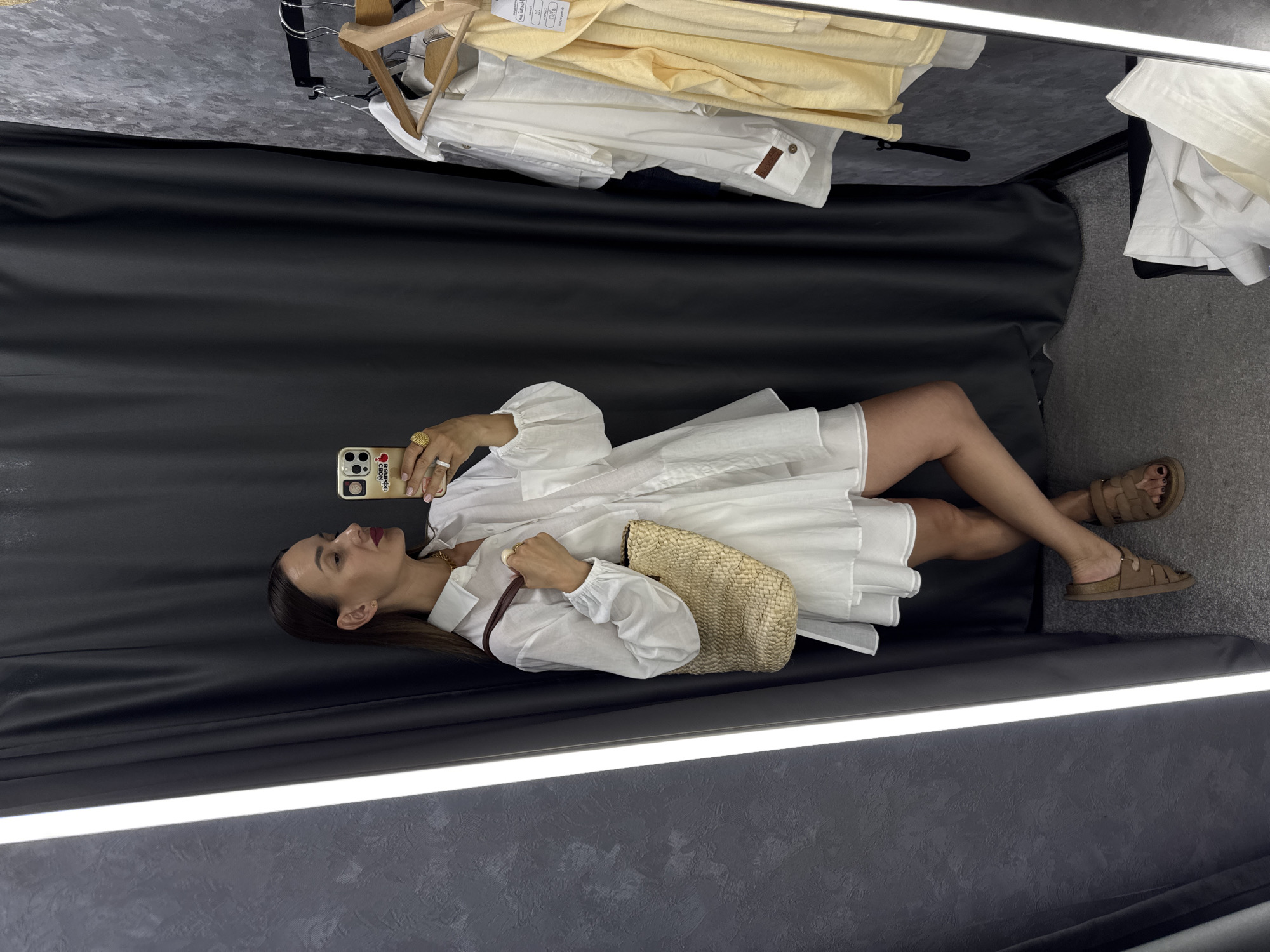 A young woman stands in a changing room, taking a mirror selfie with her phone. She is wearing a white shirt dress with ruffled layers and brown sandals. She carries a straw tote bag over her shoulder. Clothing hangs on a rack to her right, and a full-length mirror is to her left, illuminated by a bright strip light. The walls of the changing room are textured gray, and the floor is carpeted in gray. The woman has long dark hair pulled back from her face, and she is wearing red lipstick and jewelry.