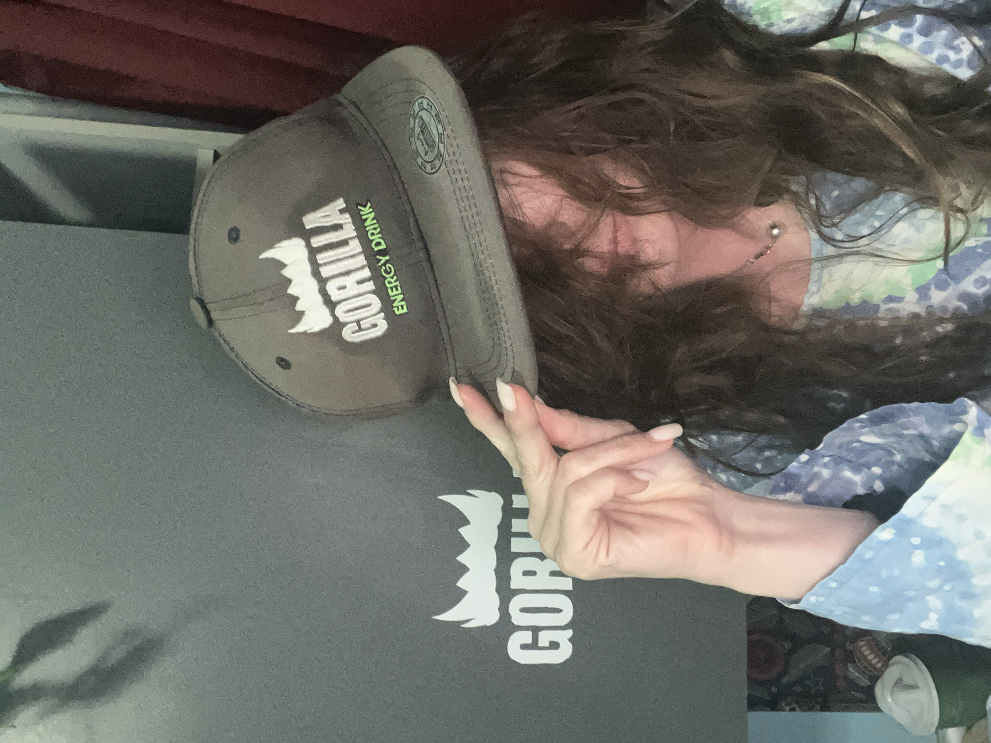 A person is holding a gray baseball cap with the "Gorilla Energy Drink" logo, posing in an indoor setting. The logo is white with green lettering. The person's long brown hair partially obscures their face, and they are wearing a blue and white patterned top. Their hand with manicured nails is visible. The background includes a gray surface with a larger version of the "Gorilla" logo, and a maroon curtain. The location is not identifiable from the image.