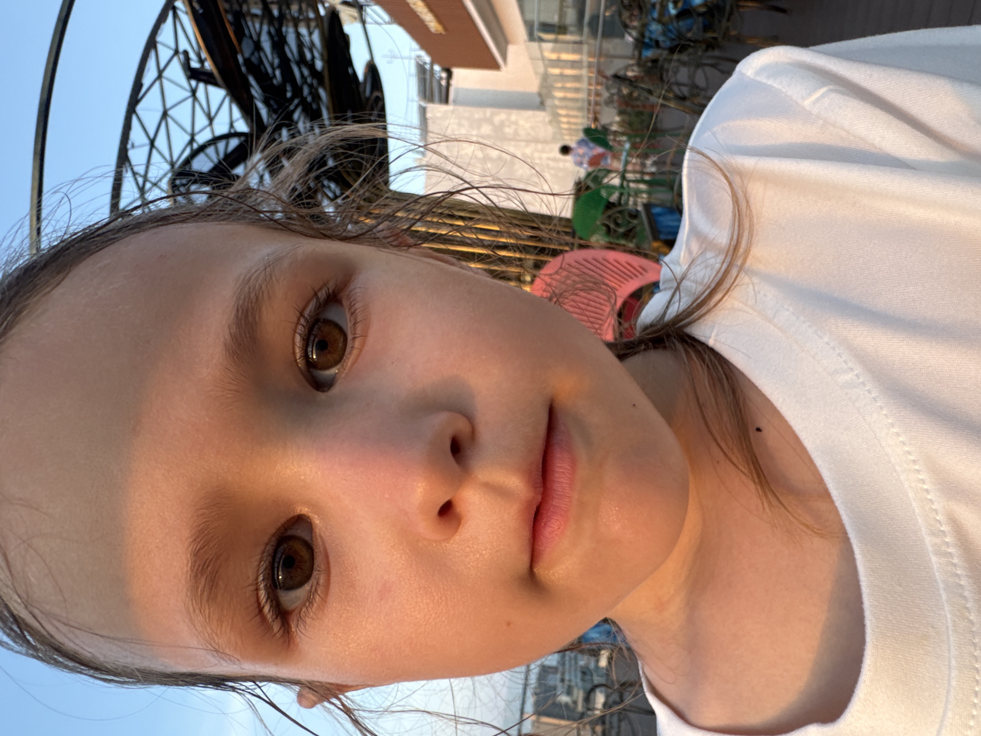A close-up photograph features a young child, centered, looking directly at the camera with a neutral expression. The child has fair skin with a strong golden-orange illumination on the left side of the face and forehead, indicative of directional, warm sunlight. She has brown eyes, a small nose, and closed lips. Her brown hair, appearing damp, is pulled back, with loose strands framing her face, particularly on the right. She wears a plain white crew-neck t-shirt. A small dark mole is visible on her lower neck.
The background depicts an outdoor location under a clear light blue sky. To the right and behind the child, a large, dark, open-framework geodesic-style lattice structure is prominent. Further back, multi-story buildings are visible, one featuring a rectangular sign with dark text on an orange-brown background, partially reading "NHA..." Below these structures, an outdoor seating area contains multiple tables and chairs, some blue and some green. A distant figure in a patterned shirt is observed within this area. A pink comb is partially visible in the lower right. The ground surface appears to be wooden decking. This scene is located in Nha Trang Ward, Vietnam.
