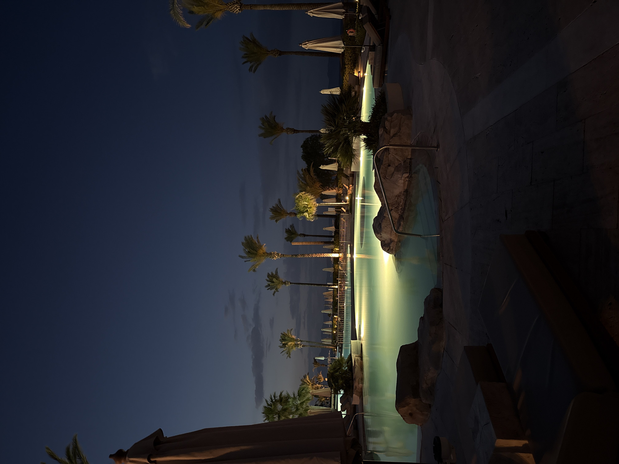 At night, a long swimming pool is illuminated, casting a bright glow across the water. Palm trees line the pool's edge, silhouetted against the dark sky. A row of white parasols stands next to lounge chairs. A dark stone barrier lines the pool. The image is rotated counterclockwise. Based on the context, the location is likely the city of Kos, Greece.