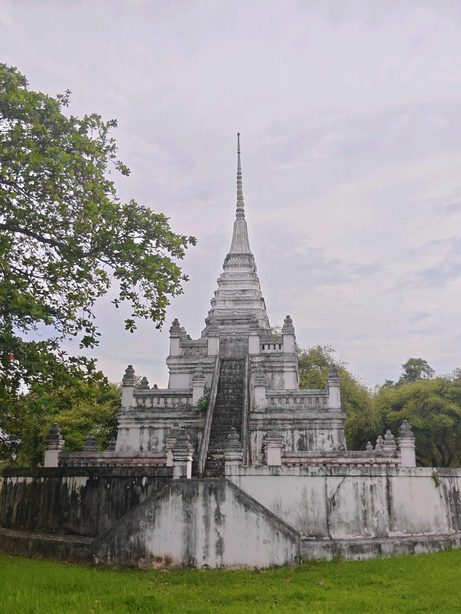 The image shows a white stupa in the city of Phra Nakhon Si Ayutthaya City Municipality, Thailand. The stupa is surrounded by lush green trees and is set against a cloudy sky. The stupa is a tall, multi-tiered structure with a pointed spire at the top. It is made of white stone and is decorated with intricate carvings. There are stairs leading up to the stupa, and a wall surrounds the base. The grass in front of the stupa is green and well-maintained. The scene appears serene and peaceful.