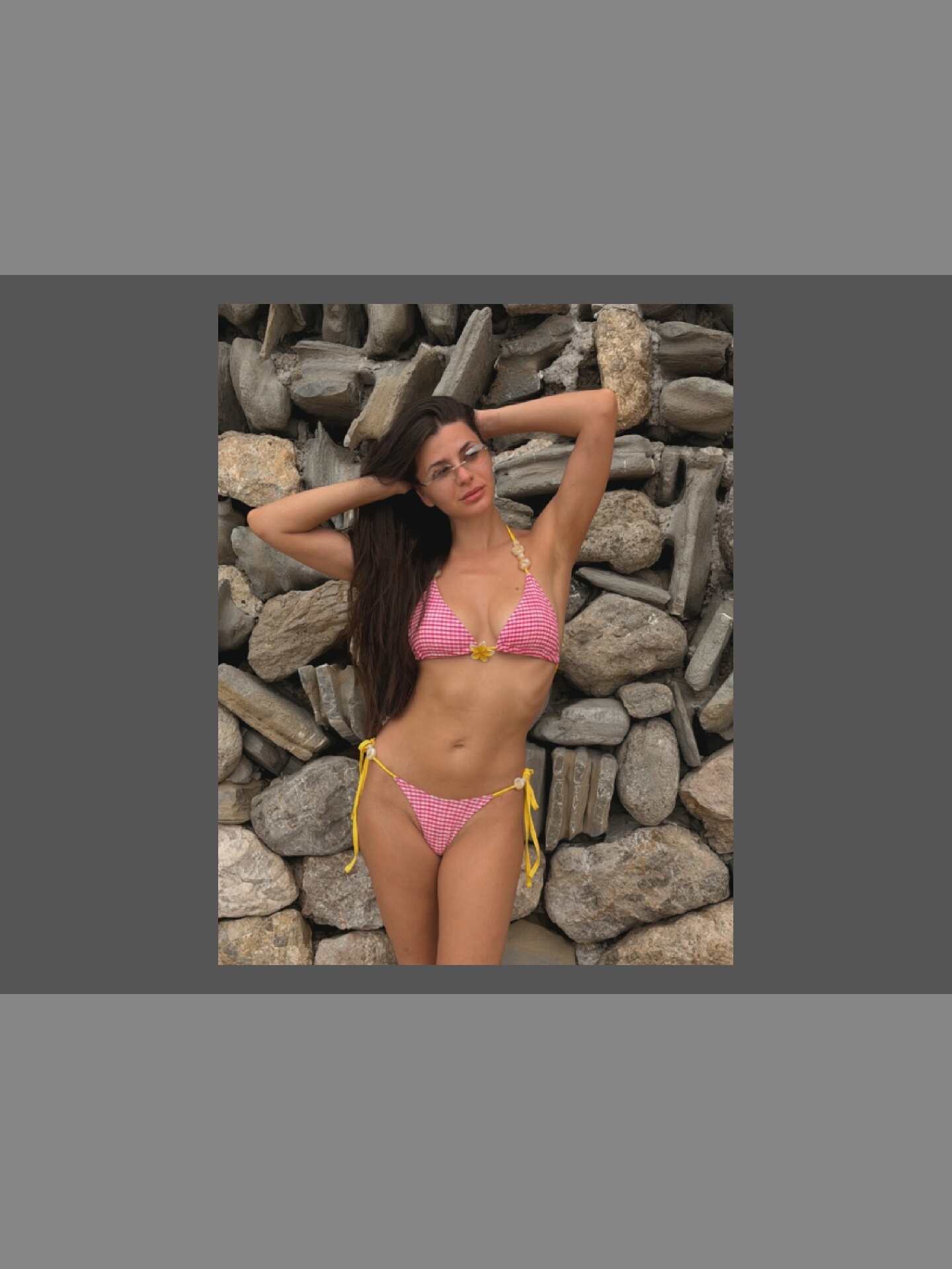 A full-shot, medium-angle, vertical photograph shows a woman posing in a pink and white gingham bikini against a stone wall. The woman has long, dark hair that falls over her shoulders and is wearing glasses. Her arms are behind her head, and her body is angled towards the viewer. The bikini is tied with yellow strings, and the top has a small yellow flower embellishment. The stone wall behind her is rough and textured, made of various sized gray and brown stones. The lighting suggests it is daytime, possibly outdoors. The woman's expression is neutral, and the overall mood is relaxed and casual. There is no visible text in the image.