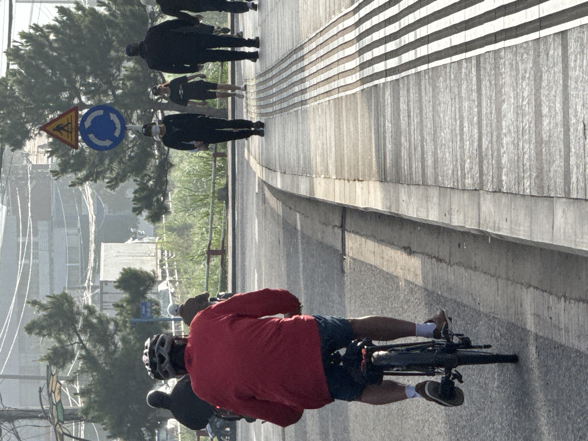 In Phường Xuân Hương - Đà Lạt, Vietnam, on a paved road with a textured pedestrian walkway to the right, a person wearing a red long-sleeved shirt, blue shorts, white socks, and brown shoes is cycling away from the camera. The cyclist is wearing a black helmet with a gray design. Several other pedestrians are visible in the background walking on the pedestrian walkway. One pedestrian, a person wearing a black mask, black clothing, and a white baseball cap, is walking to the left. Further down the walkway, another person in black shorts and a black t-shirt with a white graphic is walking. Behind them, a person in a black jacket and dark pants is walking, followed by another person in similar dark attire. Traffic signs, including a yellow triangular warning sign with a pedestrian symbol and a blue circular sign with white arrows indicating a roundabout, are mounted on a pole on the left side of the road. Trees and various structures, including what appears to be a white building or trailer and some overhead wires, are visible in the hazy background. The road has a clear lane marking.