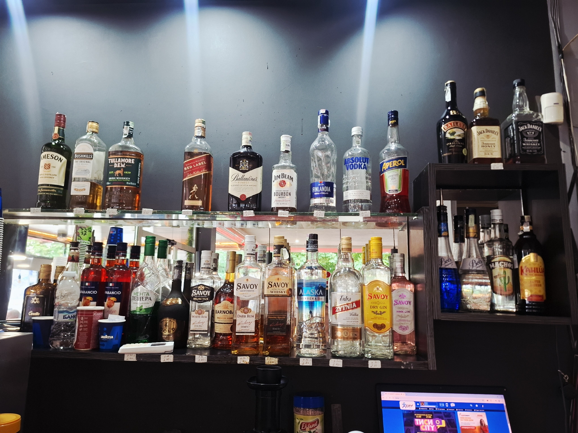 An indoor, well-lit scene showcases a wide array of alcoholic beverages displayed on two shelves, likely in a bar or store in Mezdra, Bulgaria. No people are visible.
The top shelf features numerous well-known international liquor brands. From left to right, these include Jameson, Bushmills, Tullamore Dew Irish Whiskey, Johnnie Walker Red Label, Ballantine's, Jim Beam Bourbon Whiskey, Finlandia Vodka, Absolut Vodka, and Aperol Aperitivo. Some of these bottles, like Finlandia, Absolut, and Aperol, appear to be partially empty. On a smaller, elevated shelf to the right, Baileys, Jack Daniel's Honey, and Jack Daniel's Tennessee Whiskey are displayed. Small price tags, mostly ranging from 2.20 to 4.50 (likely Bulgarian Lev), are visible below many of the bottles.
The lower shelf, reflected in a mirrored background, holds a more diverse selection of bottles, including several regional or local brands, often under the "Savoy" label. These include Savoy White Rum, Savoy Whisky, Savoy Dark Rum, Savoy Pink Gin, and Savoy Dry Gin. Other spirits like Kärnoba Premium Brandy, Alaska Vodka, Zytnia Vodka, and Menta Pestera (a Bulgarian mint liqueur) are present. There are also multiple red bottles of Parancio, Conto, and a bottle of Black Sea Gold brandy. Practical items like a plastic bottle of Bankia water and two red Lavazza cups are also seen. On a separate, darker shelf to the far right, Bols liqueurs (blue and clear), Machon, and Kahlua are displayed. Similar price tags are distributed across this shelf.
Below the shelves, a black bar counter is visible, with a container of "Ekland" coffee or tea powder. To the bottom right, a laptop is open, displaying a website with "CITY" and Bulgarian text "ТИСИ О CITY". The overall setting suggests a commercial establishment like a pub, cafe, or liquor store.