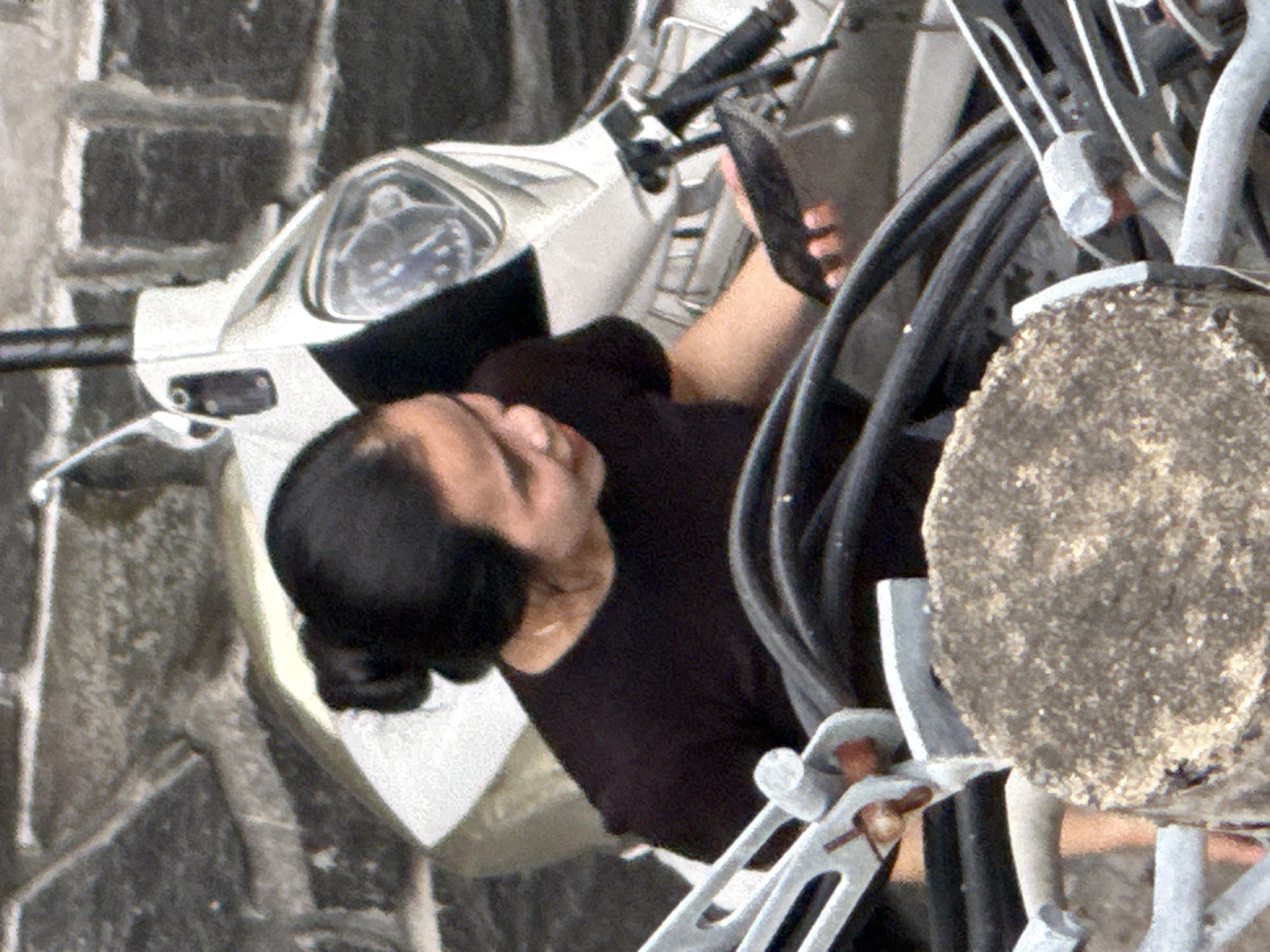 A woman is shown from the chest up, with her head turned to her left, looking down. She has dark hair pulled back and is wearing a black t-shirt. She is holding a black object in her right hand, near the handlebars of a white and silver scooter. The scooter's handlebars and some of its body are visible, along with thick black cables or hoses. The background appears to be a stone wall and a portion of the scooter's front end. The image is a close-up, and the scene is likely outdoors, possibly in a city like Nha Trang, Vietnam, given the presence of a scooter and the architectural style of the background.