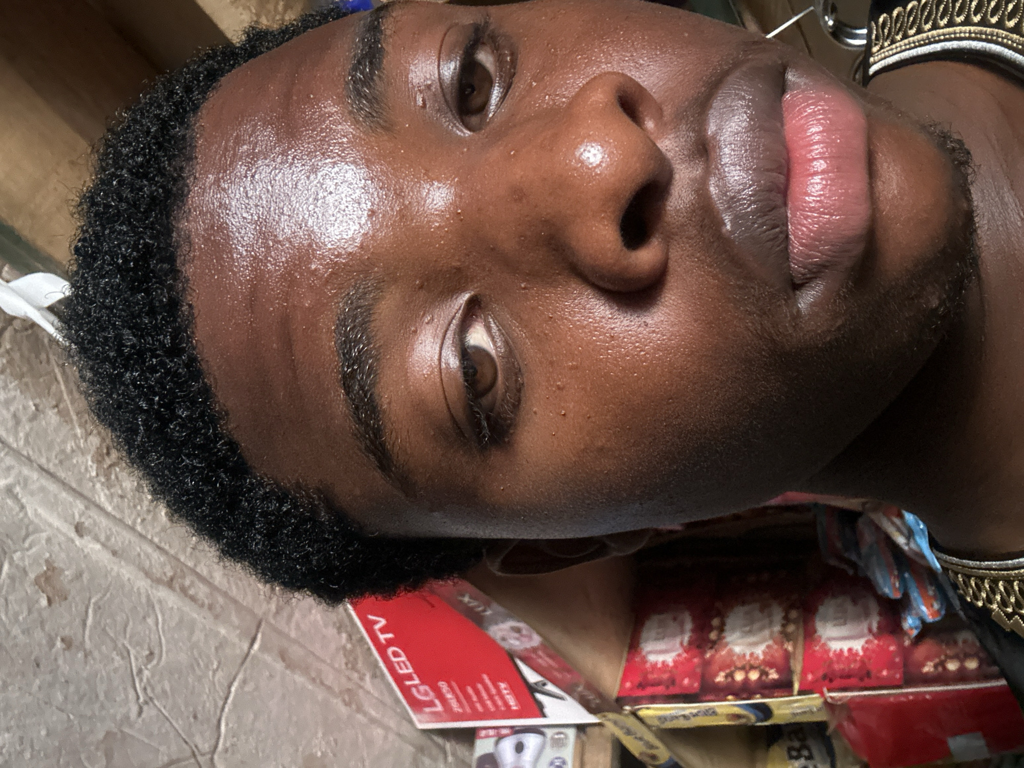 Close-up of a Black man's face. His skin is dark and shiny, possibly due to sweat or oil. He has short, curly black hair. His eyes are dark brown, and his lips appear pink and full.
The background shows items on a surface. There is a red box with "LG LED TV" printed on it, other boxes are also visible. The image was taken in Bukuru, Nigeria.