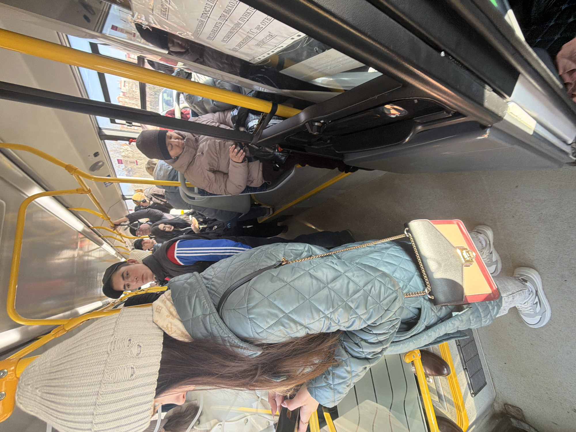 The scene is inside a bus in Krasnoyarsk, Russia, with several people present. A woman wearing a light blue quilted jacket, a white knitted hat, grey jeans, and white sneakers is in the foreground. She's holding a black smartphone and has a shoulder bag with a gold chain. A young man in a black hat and a blue and red striped jacket is partially visible. Behind them, there are more people seated. Above, near the bus ceiling, is an information sign written in Russian. The interior of the bus has yellow handrails. The image is taken from a slightly skewed perspective.
