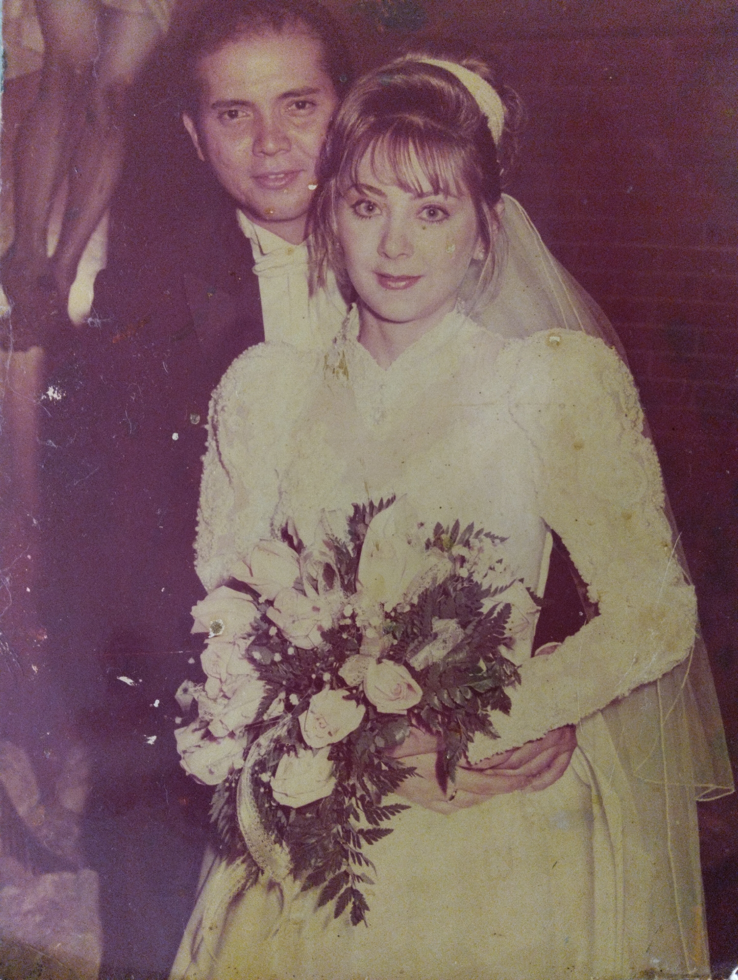 A photograph depicts a man and a woman, likely at their wedding. The man, on the left, is wearing a dark suit with a white bow tie. The woman, in the center, is wearing a long-sleeved white wedding dress and a veil. She is holding a bouquet of flowers. A man's hand is visible, holding the woman around her waist. The background is indistinct but appears to be a brick wall. The location is Unknown, Unknown.