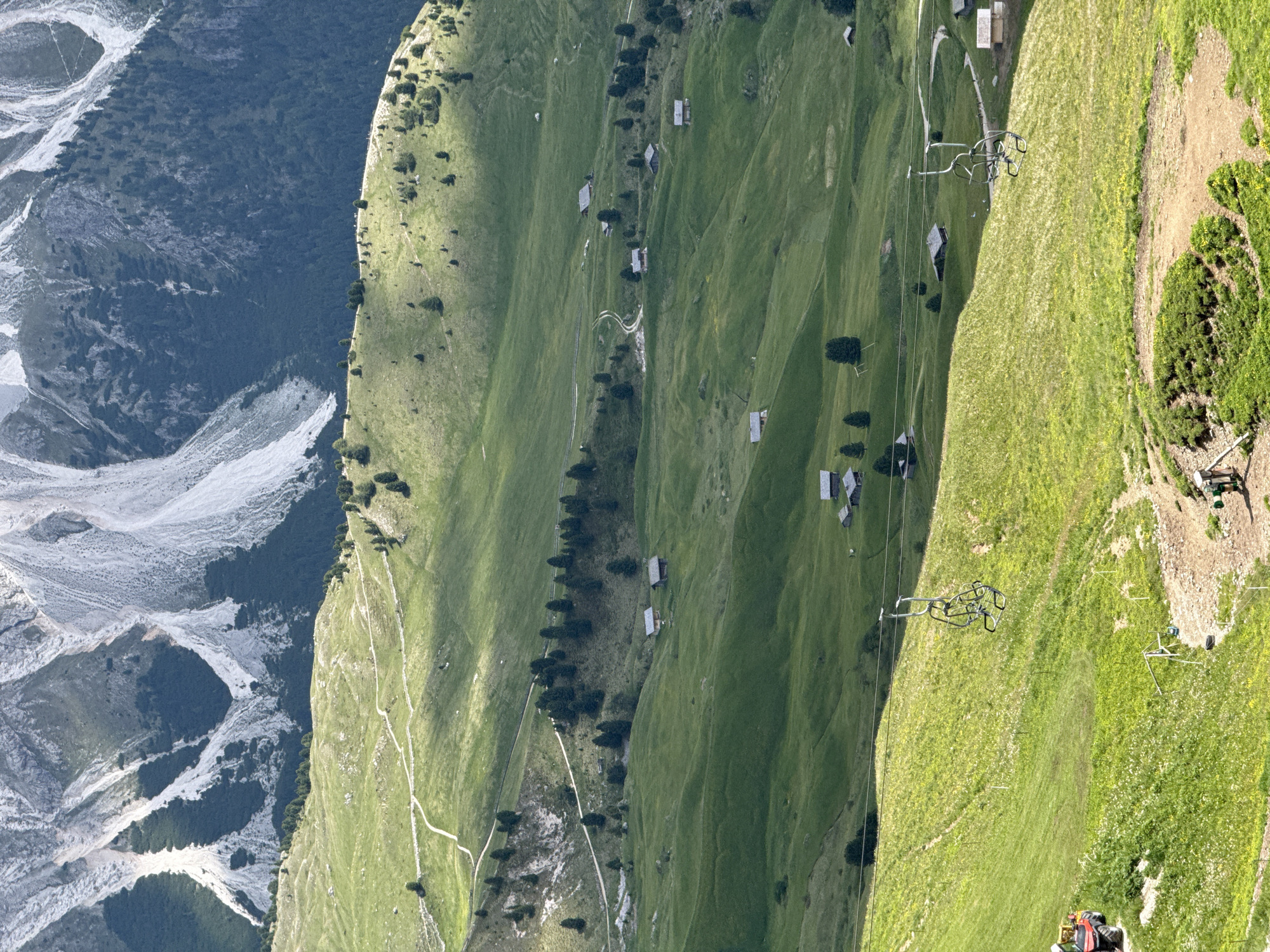 This is a vertical, high-angle shot of a mountainous landscape in Santa Cristina Gherdëina - St. Christina in Gröden - Santa Cristina Valgardena, Italy. The foreground features a bright green, grassy slope with scattered wildflowers. A red tractor is partially visible on the left. A ski lift chair hangs suspended over the slope. Further back, rolling green hills are dotted with small wooden cabins and scattered trees. A winding dirt path is visible traversing the hills. In the background, steep, rocky mountain faces with patches of dark green trees rise dramatically. The sky is not visible. The lighting suggests a bright, sunny day, with shadows cast across the terrain.