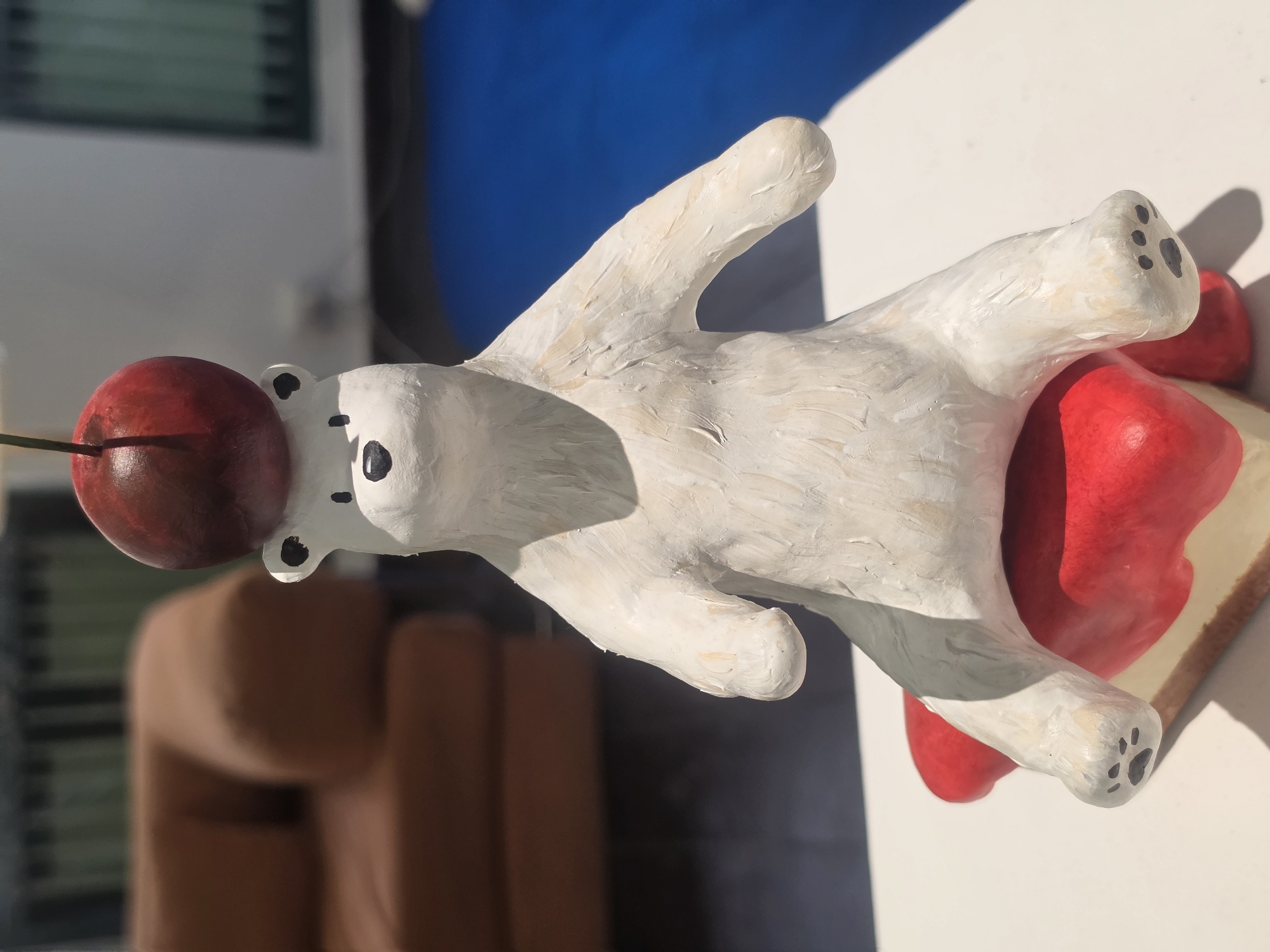 The image shows a white, possibly ceramic or plaster, polar bear figurine lying on its back. A large, red cherry rests on its forehead, seemingly balanced there. The bear is situated on what appears to be a piece of toast with a thick layer of red jam or icing. The paws of the bear have black spots representing paw pads.
The background features a blurred view of what seems to be an interior scene, possibly with a sofa and partially visible window blinds. A bright blue area is also seen in the background, possibly a painted wall or a clear sky. The location is most likely indoors, given the presence of the furniture and blinds. There's no information to confirm the scene is in Bangkok, Thailand.