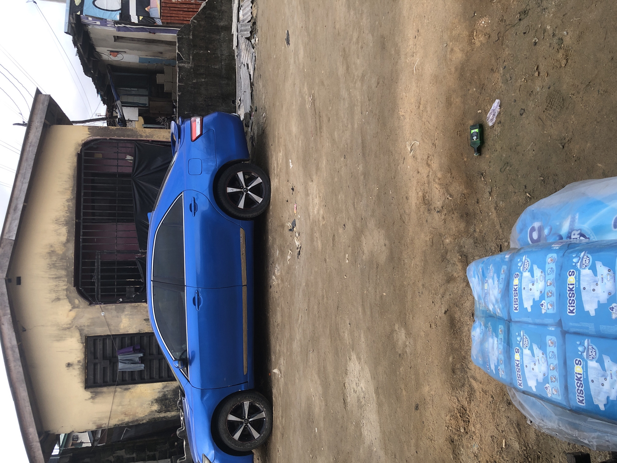In Ado, Nigeria, a blue sedan is parked on a dirt road in front of a weathered, yellow building. The building has a corrugated metal roof and a dark wooden door with laundry hanging on it. A metal gate with bars covers a window. To the right, another structure with a tin roof and a rough concrete wall is visible. In the foreground, a stack of blue diaper packages is wrapped in plastic. Further to the right on the dirt road, a green bottle lies on its side, accompanied by a crumpled piece of plastic. The ground is uneven and littered with debris.