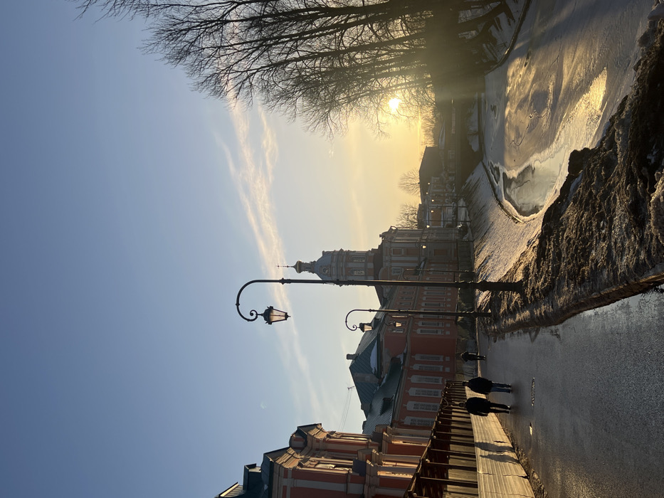 This image depicts a winter urban scene in Saint Petersburg, Russia, specifically along the Monastyrka River adjacent to the Alexander Nevsky Lavra complex. The scene is illuminated by low-angle sunlight, indicative of either sunrise or sunset.
The upper two-thirds of the frame is dominated by a clear, light blue sky transitioning to a golden-orange hue near the horizon on the right, where the sun is visible, partially obscured by trees. Wispy white clouds streak across the mid-left sky.
In the foreground-left, a wet, dark pavement or road extends diagonally into the distance. Three individuals are observed on this pavement; two men in dark winter clothing walk side-by-side in the mid-ground, moving away from the viewer, while a third, smaller figure is further in the background. Adjacent to the pavement on the left are multi-story buildings with salmon-pink facades and white architectural details, likely part of the Lavra complex. The closest building features a temporary wooden barrier or hoarding along its lower section. Two ornate black streetlights with illuminated yellow lanterns line the pavement, one prominently in the mid-ground and another further back.
To the right of the pavement and streetlights, the Monastyrka River flows, partially frozen with irregular patches of ice and dark open water reflecting the golden sunlight. The riverbanks are covered in dirty snow and slush. A distinct historical building, identified as the Church of St. John the Baptist at the Alexander Nevsky Lavra, stands prominently in the mid-ground on the far bank of the river. It features a reddish-orange lower section with white classical elements, transitioning to a lighter-colored octagonal drum supporting a domed roof and a tall spire topped with a cross. Further down the river in the background, a low bridge spans the waterway. A dense cluster of bare deciduous trees lines the right bank of the river, their silhouettes stark against the bright sky and setting sun.