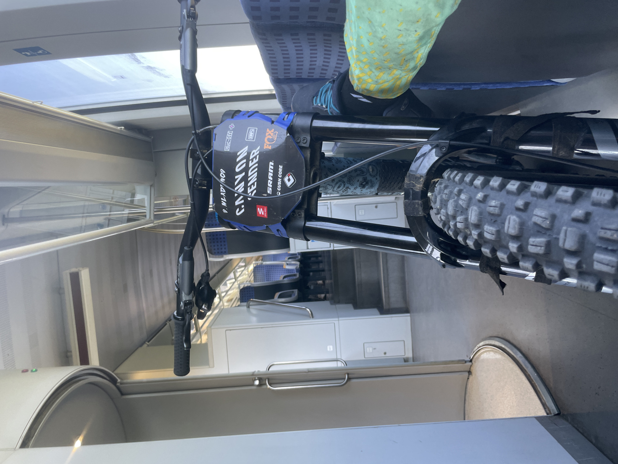 A first-person perspective shot from inside a train carriage shows a mountain bike parked vertically. The bicycle's handlebars are in the foreground, followed by the front fork and tire. A black and blue number plate is attached to the handlebars, displaying "CANYON ENDER" and several sponsor logos, including SRAM and FOX. The train carriage has blue and gray seats visible in the background, along with a window. The floor is gray, and to the left, a curved doorway with a silver handle is visible. The environment suggests the bicycle is being transported by train, possibly to or from Merichleri, Bulgaria, given the context. There are no people clearly visible in the frame, only the presence of the bicycle and the interior of the train.
