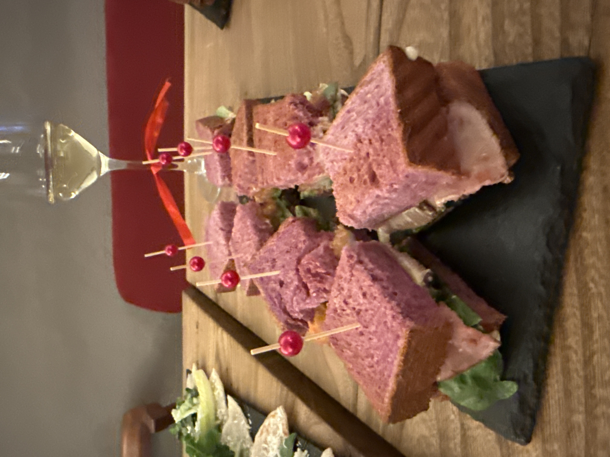 Here's a detailed description of the image:
**General Description:** The image depicts a close-up shot of food items served on a wooden surface. There is a plate of triangular sandwiches, with a glass of white wine on the side. The sandwiches are arranged on a dark slate-like plate.
**Subjects and Objects:**
* **Sandwiches:** These are notable for their pink-purple-tinged bread, secured with toothpicks topped with red beads. Lettuce leaves can be seen, indicating the filling.
* **Glass of White Wine:** A stemmed wine glass is partially visible, filled with a light-colored liquid, presumably white wine.
* **Plate:** A dark, possibly slate, plate holds the sandwiches.
* **Table:** The surface upon which the food is placed is a wooden table.
* **Ribbon:** A red ribbon is visible near the glass of wine.
* **Salad:** There is a salad bowl filled with sliced green ingredients on the left-hand side.
**Environment:**
* The setting appears to be indoors, possibly in a restaurant or dining establishment.
* The color palette is warm, with the wood tones contrasting against the food and the slate plate.
* A red chair/furniture piece is visible in the background.
**Moscow, Russia:** There are no overt visual cues (architecture, signage, etc.) to confidently state the location is Moscow, Russia. This would be speculative.