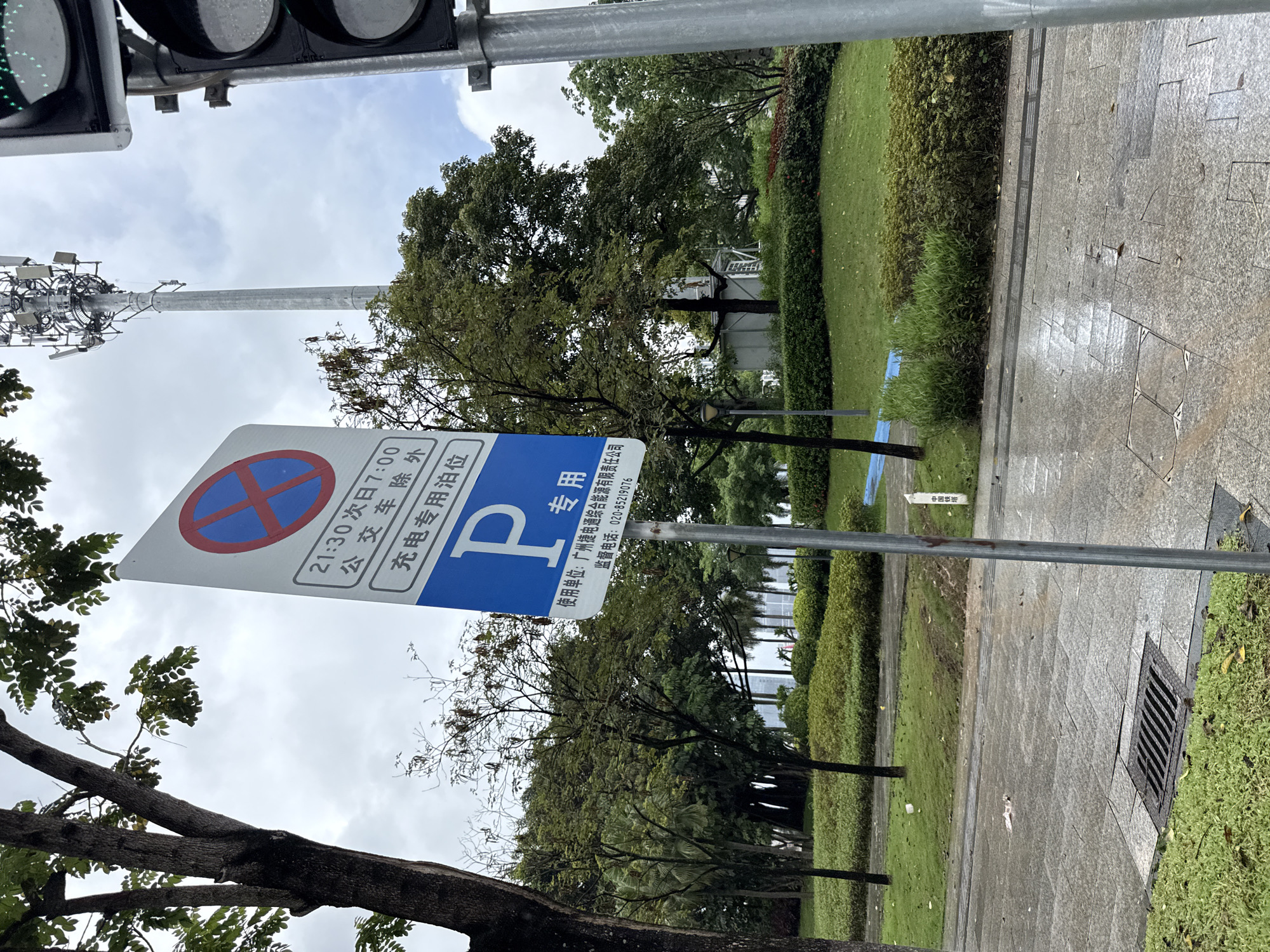 The image depicts a street scene in Haizhu District, China, likely on a cloudy or overcast day, given the diffused lighting and the wet pavement. A blue and white parking sign is prominently displayed in the foreground, indicating restricted parking times from 21:30 to 7:00, with exceptions for public transport. Below this, it specifies "Electric Vehicle Charging Parking Only". A large "P" is visible on the sign, signifying parking. Text below this provides contact information for a responsible unit, "Guangzhou Jietong Integrated Energy Co., Ltd.", and a supervisory phone number: 020-85219076.
To the right of the sign, a traffic light is partially visible, with its top green light illuminated. In the background, a tall telecommunications tower stands against the sky. The surroundings include lush green grass, manicured bushes, and trees with dense foliage. The pavement appears wet, suggesting recent rain, and a metal grate drain is visible on the left side of the road. The overall environment suggests a public area, possibly a park or a street with designated parking zones. There are no people or animals visible in the frame.
