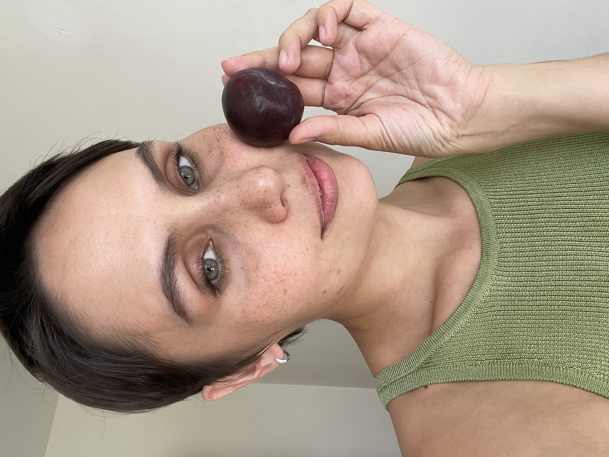 A woman is holding a plum against her cheek, near her nose. She has fair skin, freckles, dark hair, and green eyes. She is wearing a green, knitted top. She has a silver hoop earring in her left ear. The background is a plain, off-white wall. The setting is not discernable.