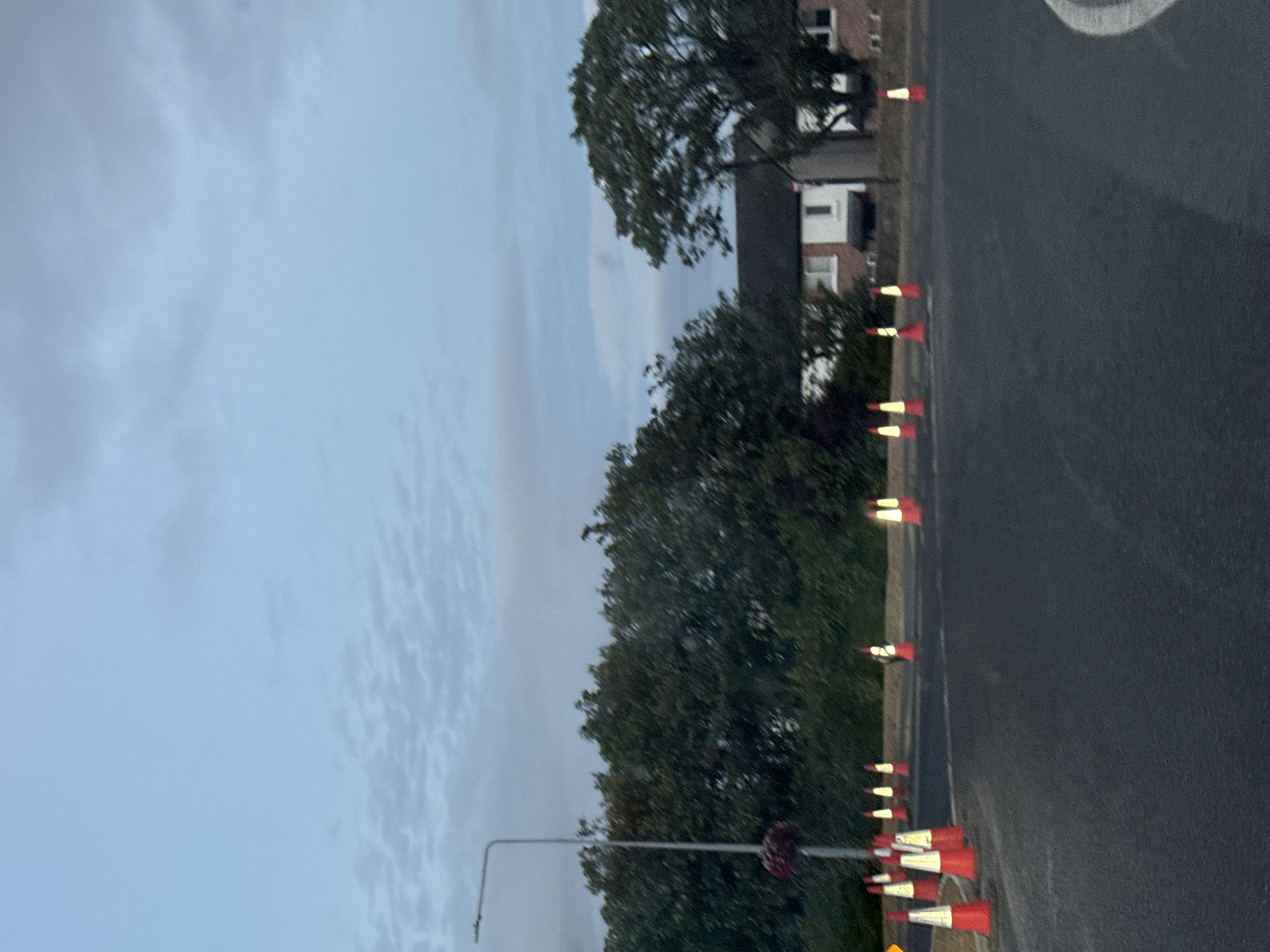 The image shows a road with several traffic cones. In the background, there are trees, houses, and a streetlamp. The sky is overcast and appears to be dusk. There are no people visible in the image. The scene is calm and quiet, with no visible activity. The location is identified as Tramore, Ireland. The traffic cones are orange and white and are placed on the side of the road. The houses in the background are brick buildings with white trim. The trees are green and leafy. The streetlamp is tall and silver. The overall impression is one of a quiet evening in a residential area.